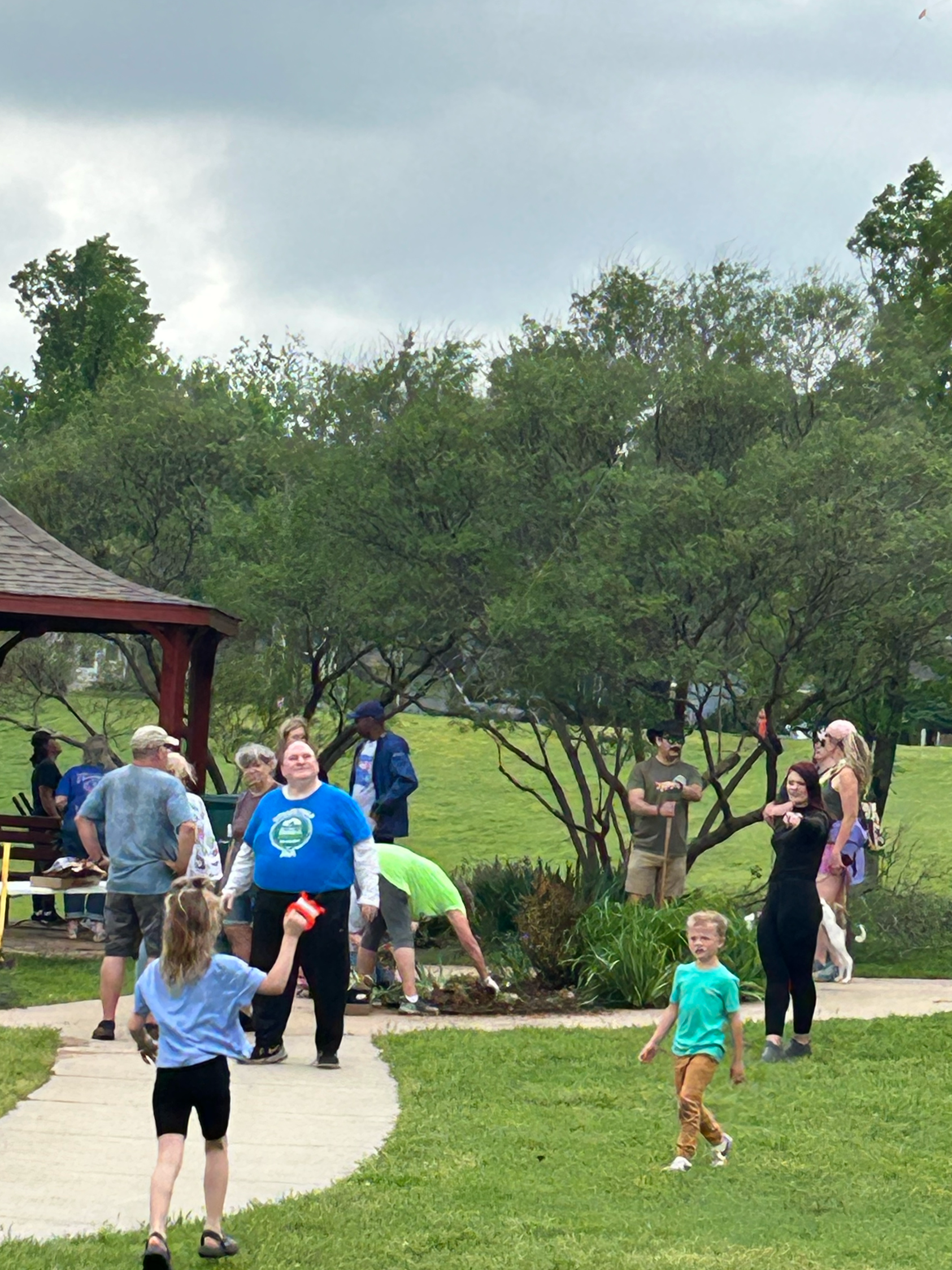 People gathering in a park near a garden bed, some planting flowers, with trees and a pavilion in the background.