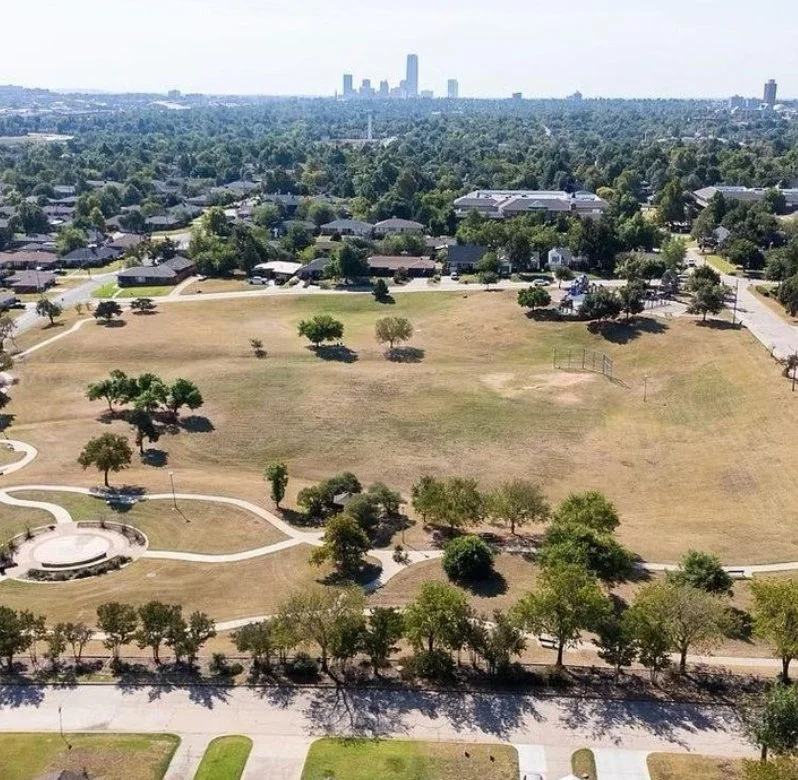 An aerial view of a park with open grassy areas, trees, paved walkways, and a small circular structure, with residential neighborhood and a city skyline in the background.