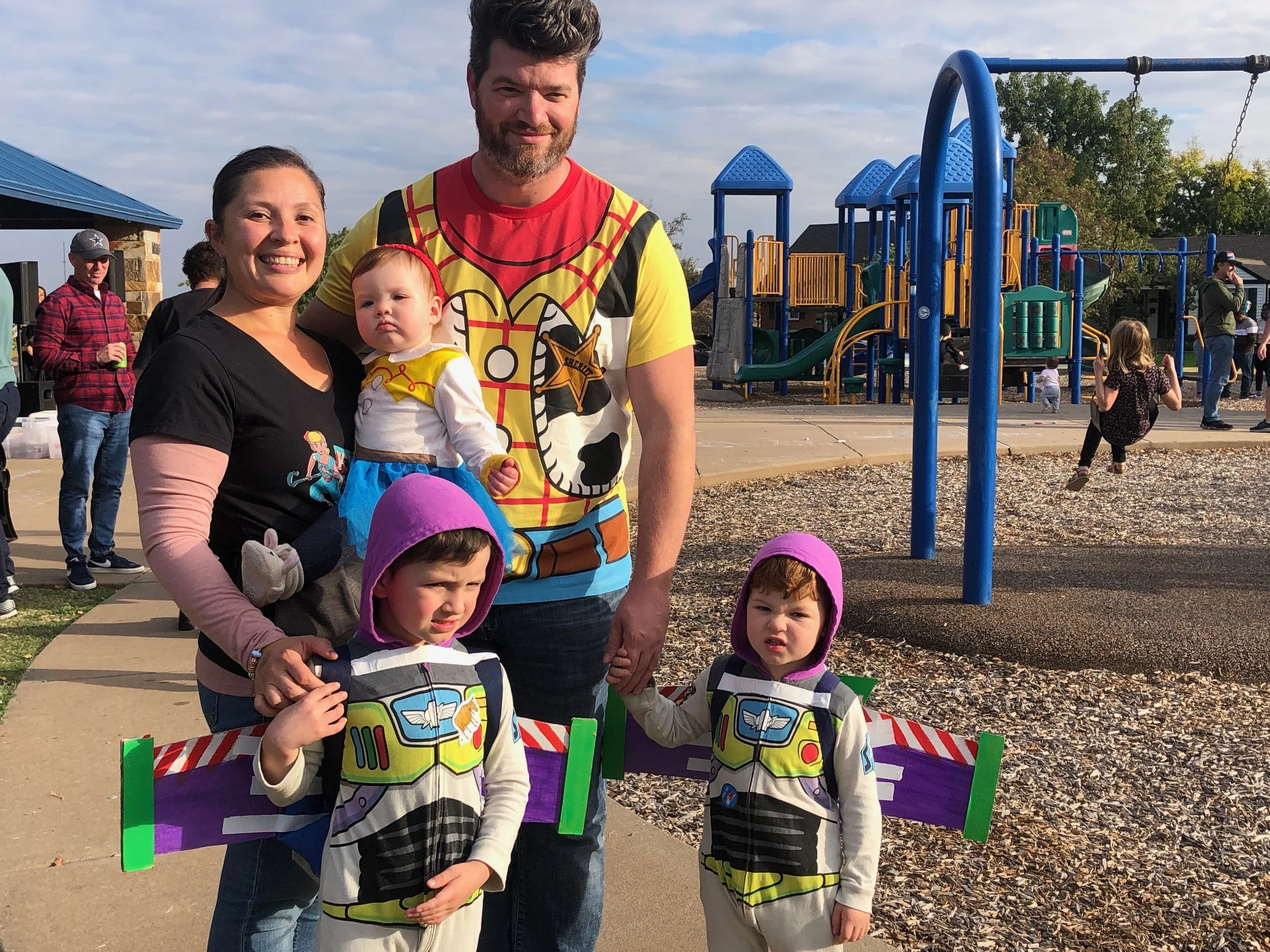 A family of five, including two young children dressed as Buzz Lightyear and two adults, posing at a playground during daytime with other people in the background.