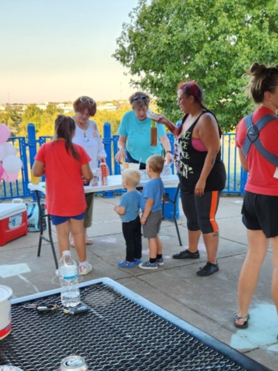 Children and adults gathered outdoors around a table with food, balloons, and drinks, on a concrete patio with a tree and a fence in the background.