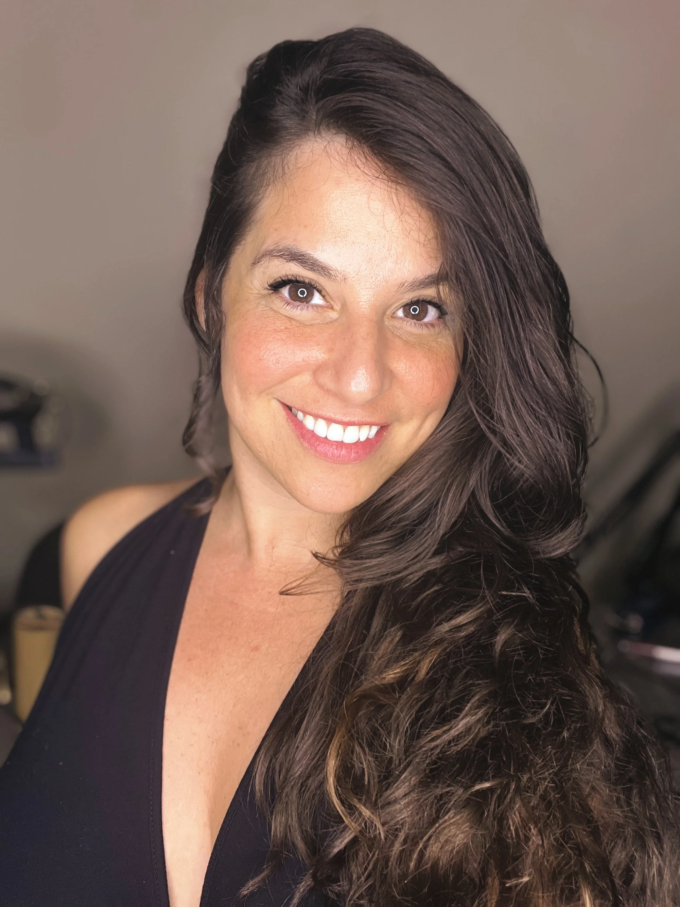 A smiling woman with long, wavy brown hair, wearing a black sleeveless top, standing indoors.