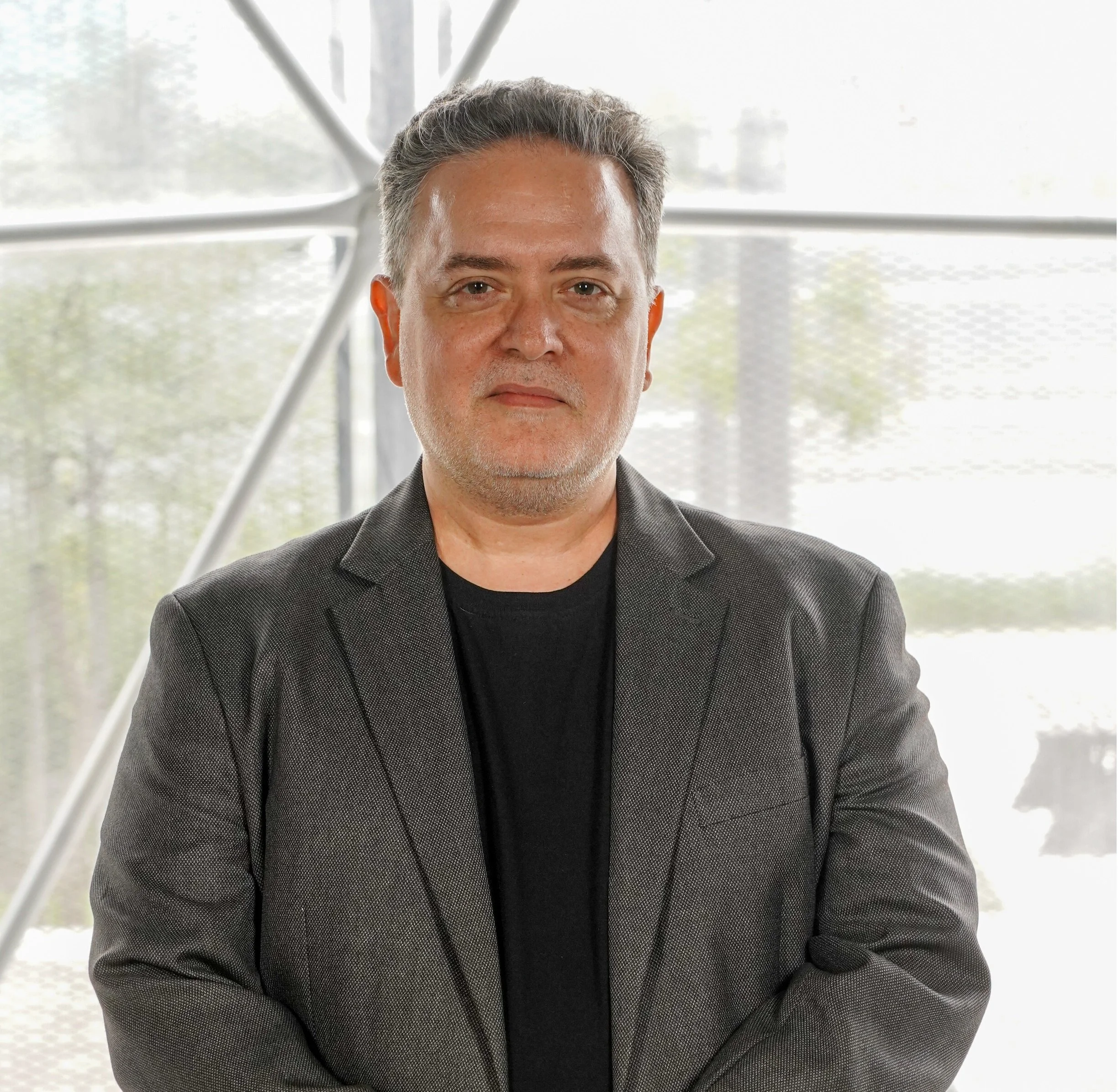 A middle-aged man with gray hair and a goatee, wearing a black blazer and black shirt, standing in front of a modern architectural background.