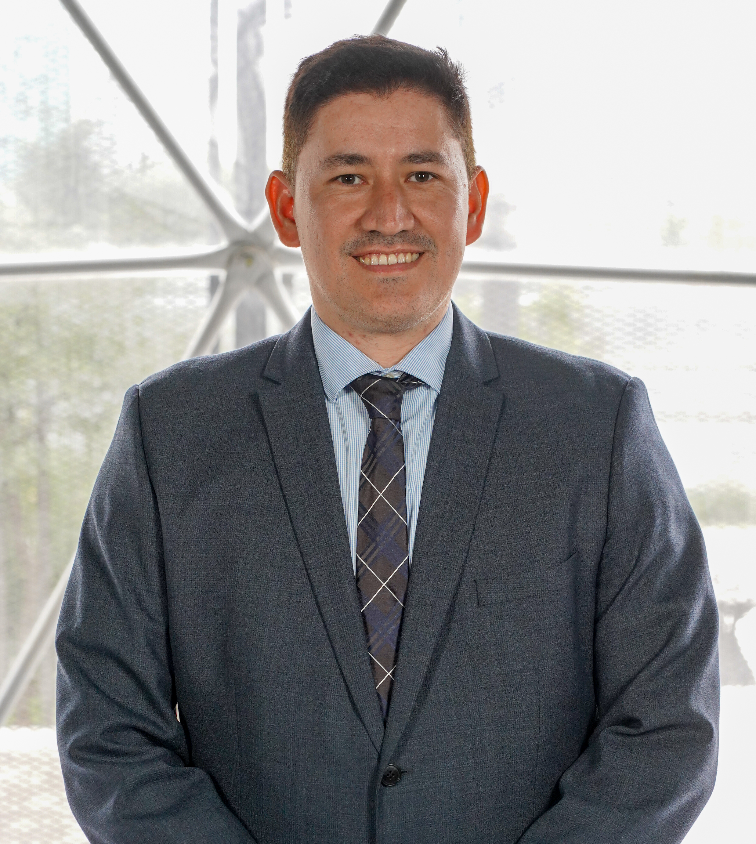 A man in a dark suit with a light blue dress shirt and a dark plaid tie, smiling, standing in front of a modern architectural background with large windows and metal framework.