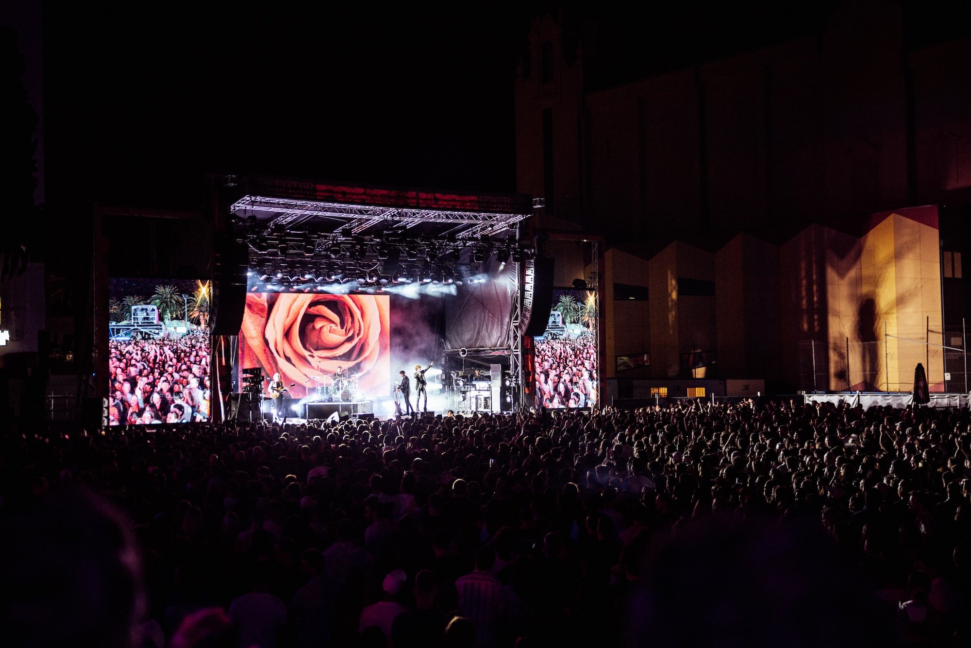 Palace Foreshore nighttime concert with a large crowd watching The Kooks perform on stage, with a large screen displaying a rose behind the band.