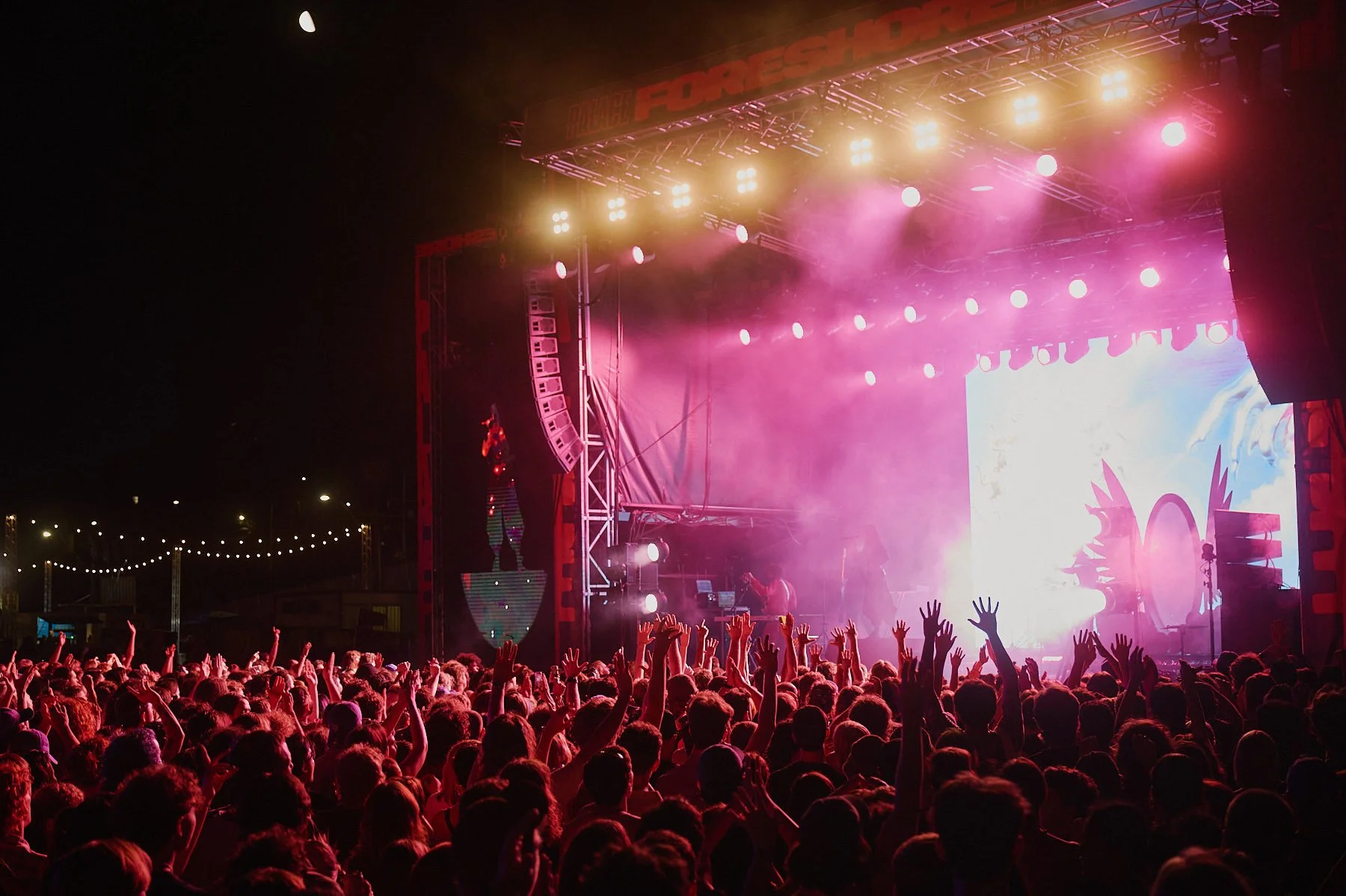 Crowd at a Palace Foreshore music concert or festival during nighttime with a brightly lit stage, pink and purple fog, and people raising their hands.