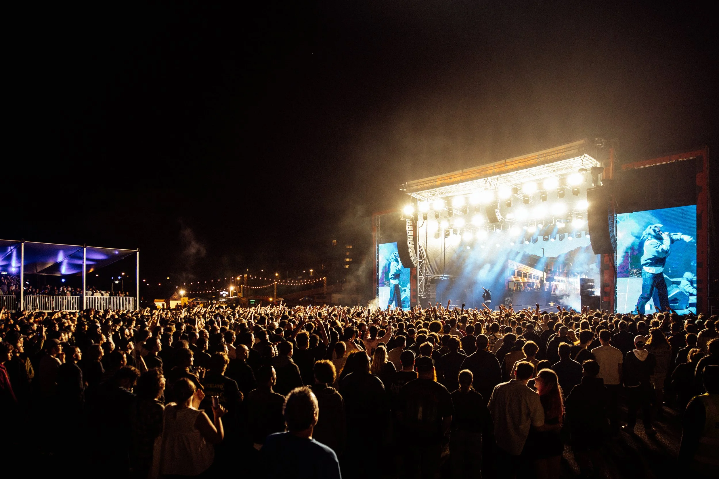 Large crowd at Palace Foreshore, an outdoor concert at night with a brightly lit stage and performers, two large screens show images of singers, and the audience is facing the stage amid colorful lights.