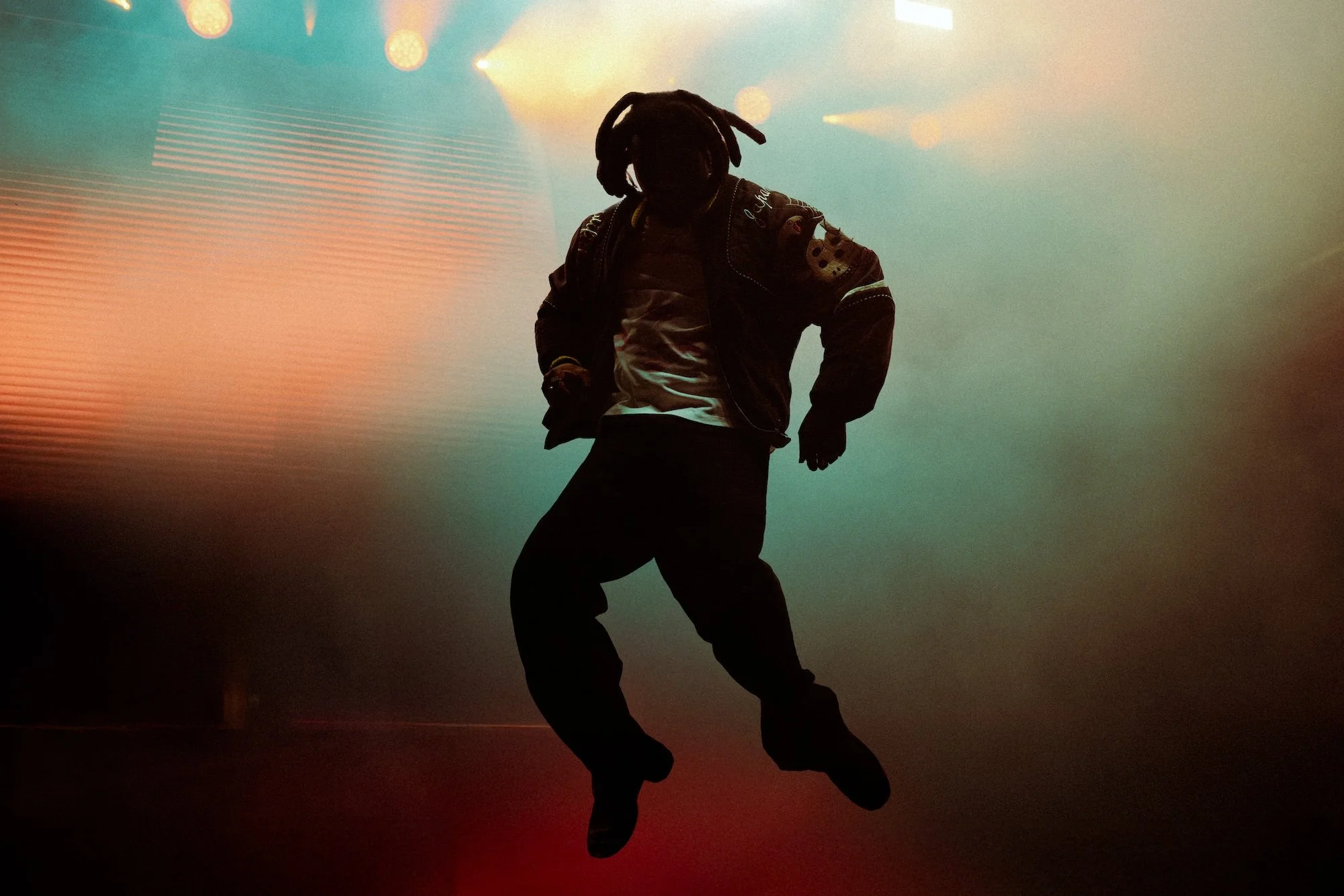A person with dreadlocks (artist, Denzel Curry performing at Palace Foreshore) jumping in the air against a colorful, hazy backdrop with stage lights.