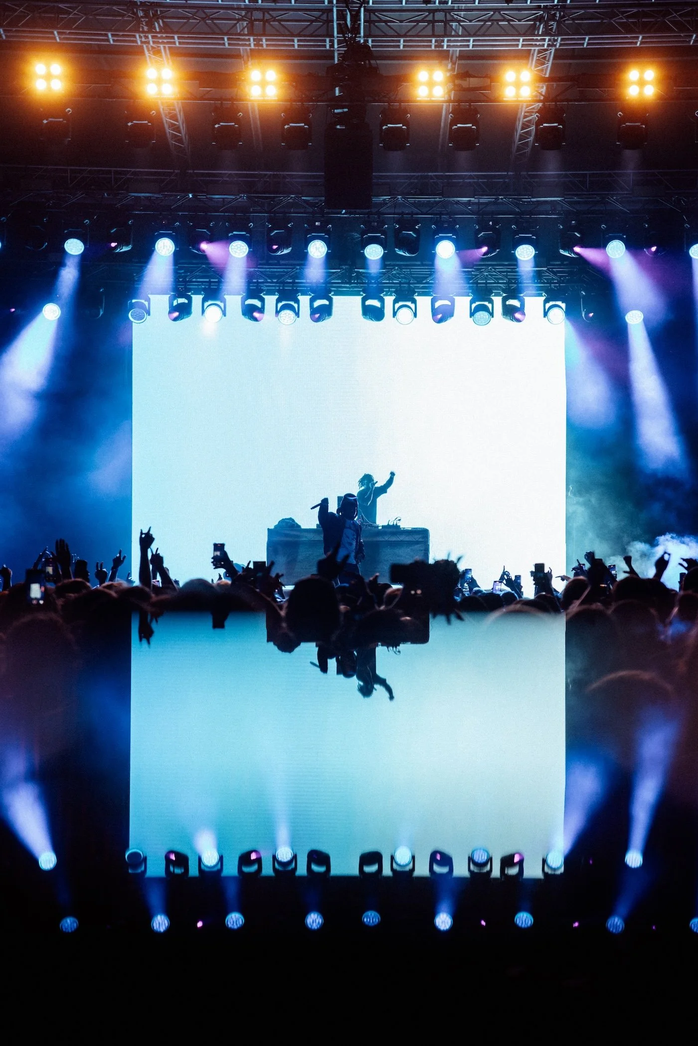 A live music concert with a DJ and Denzel Curry performing on stage at Palace Foreshore, bright stage lights, and an audience taking photos, with a large LED screen displaying the DJ and crowd in the background.