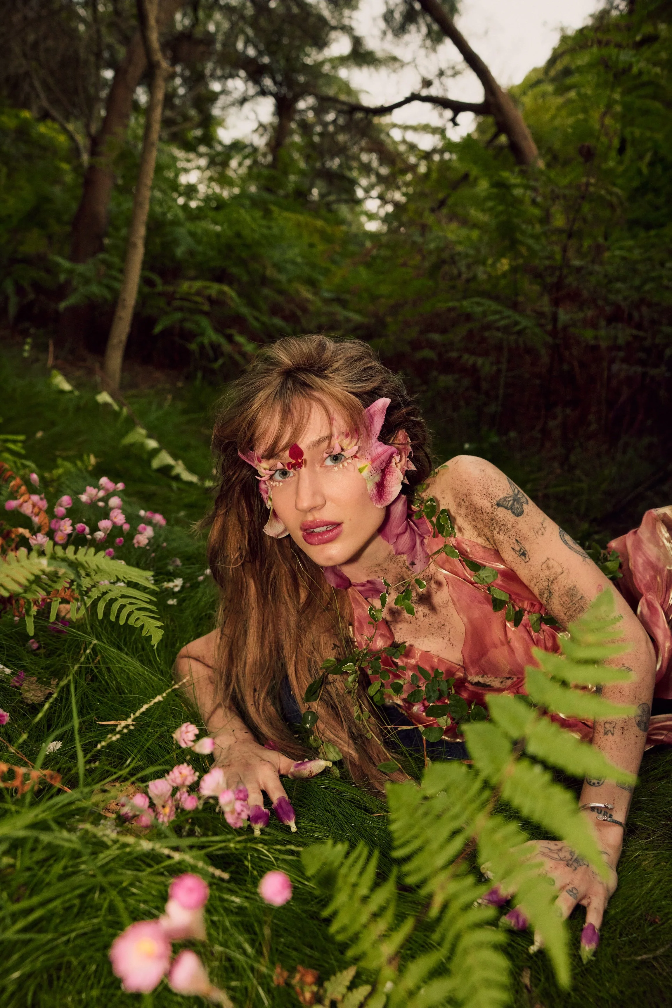A woman (Australian pop artist Peach PRC) with long, wavy red hair lying on the forest floor surrounded by pink flowers and green foliage.
