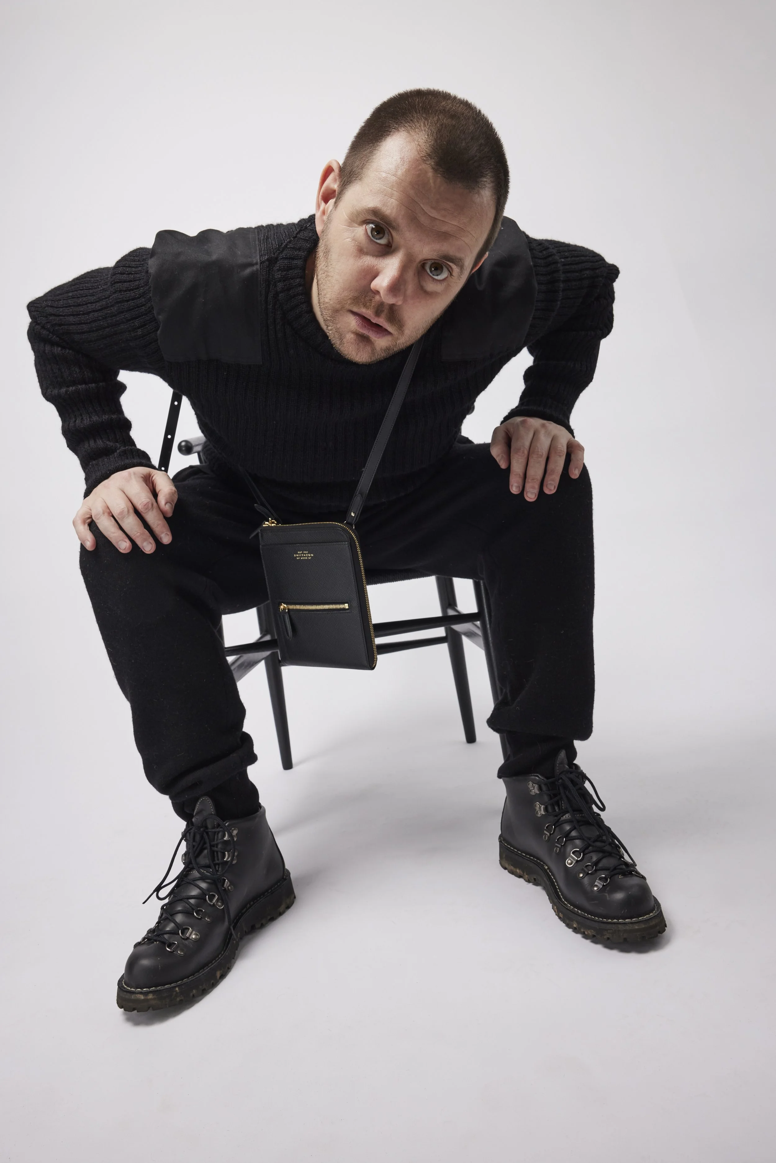 A man (Mike Skinner / The Streets) with short dark hair and a beard is squatting on a chair, leaning forward with an inquisitive expression, wearing a black sweater, black pants, and black boots, with a small black bag hanging around his neck.