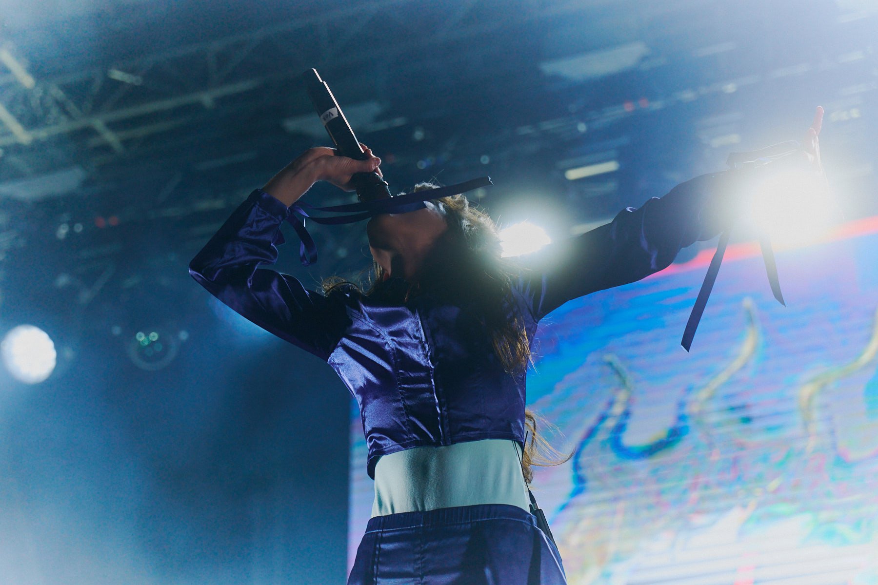 A female singer from Magdalena Bay with curly hair performing on stage with a microphone, wearing a purple satin jacket and a light-colored skirt, illuminated by bright stage lights. Playing at Palace Foreshore in 2025.