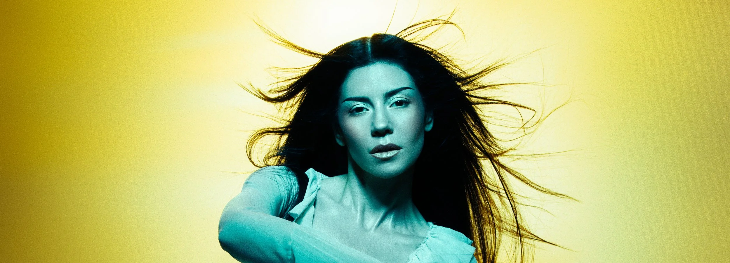 A woman, artist Marina, with long hair blowing in all directions, facing the camera with a confident expression, against a yellow background.