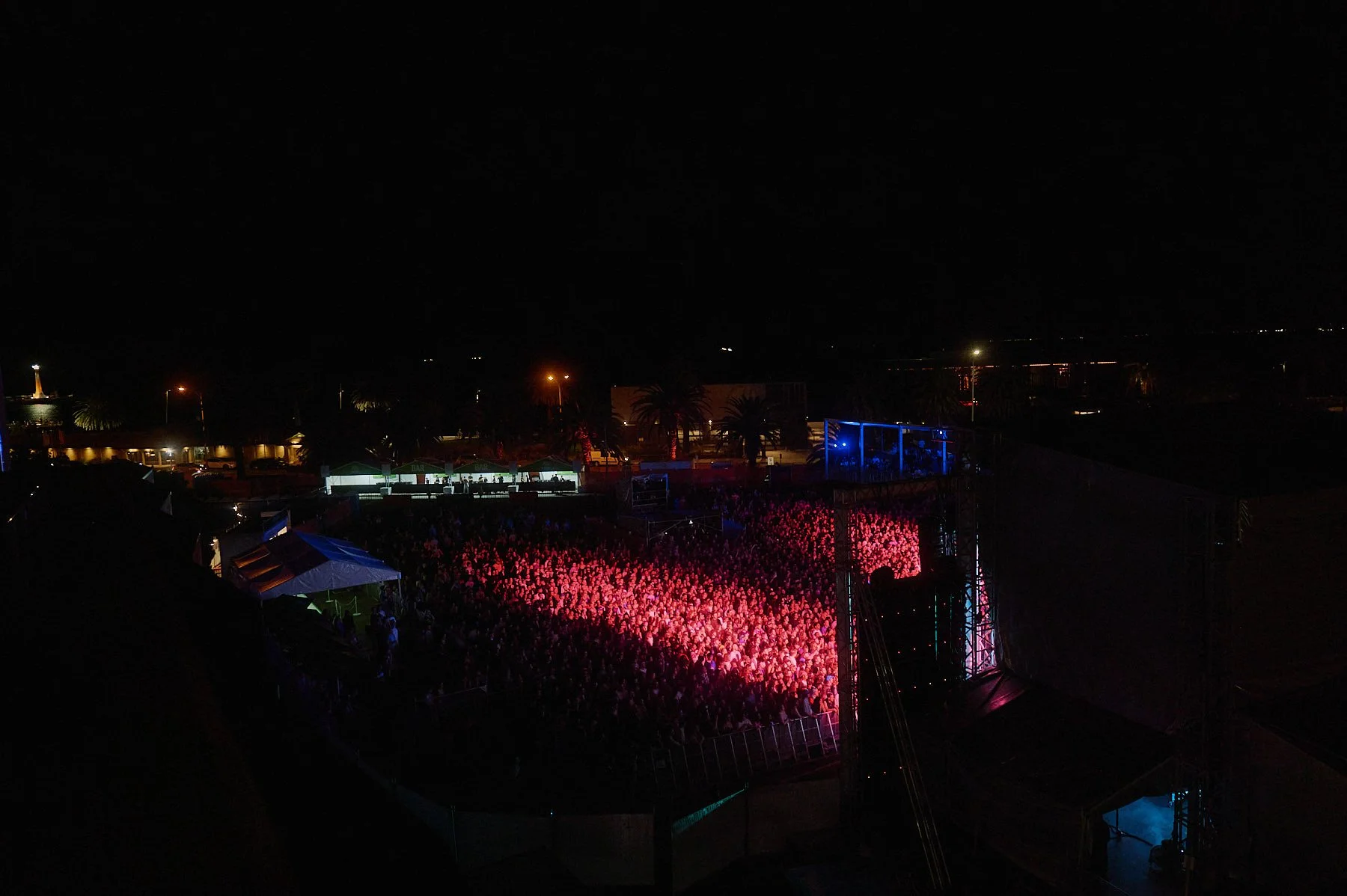 Nighttime outdoor concert at Palace Foreshore with a large crowd, stage with blue and pink lighting effects, and tents in the background.