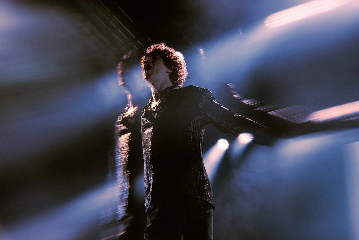 The Kooks lead singer with curly hair singing passionately on stage at Palace Foreshore, illuminated by dramatic lighting with blurred motion effects.