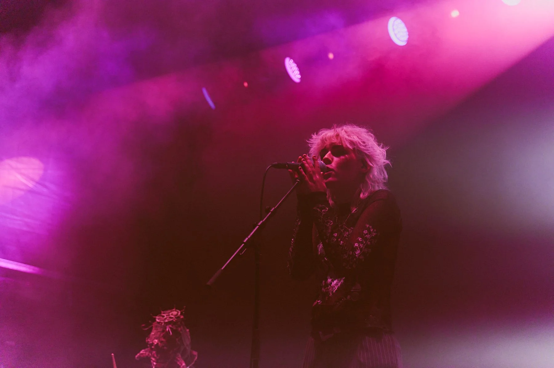 Ratbag, a female singer with blonde curly hair performing on stage at Palace Foreshore under purple and pink lights, singing into a microphone.