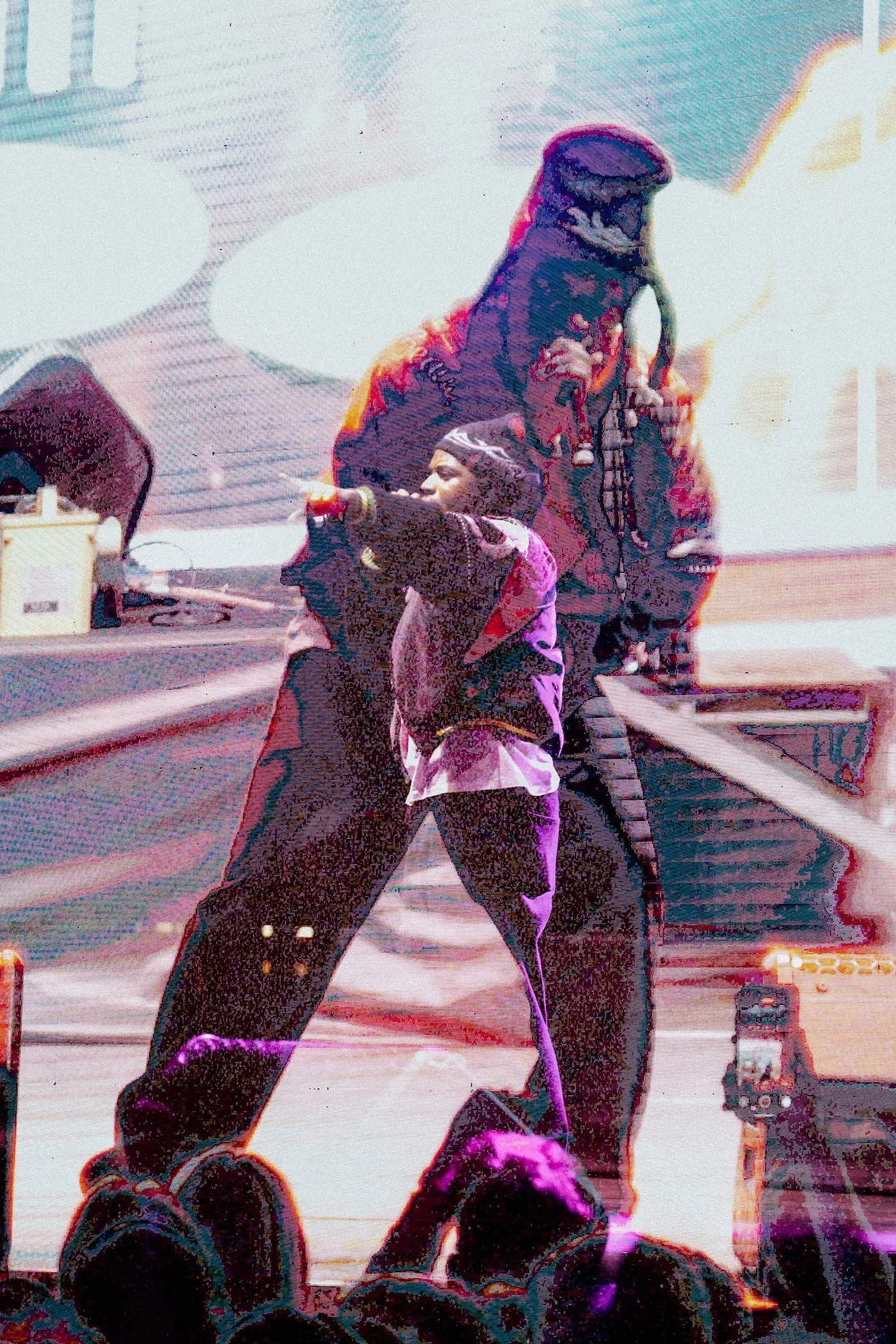 Denzel Curry at Palace Foreshore wearing a beanie and a jacket is performing on stage with a microphone. The image has a film effect and a double layer applied, making it look abstract and grainy.