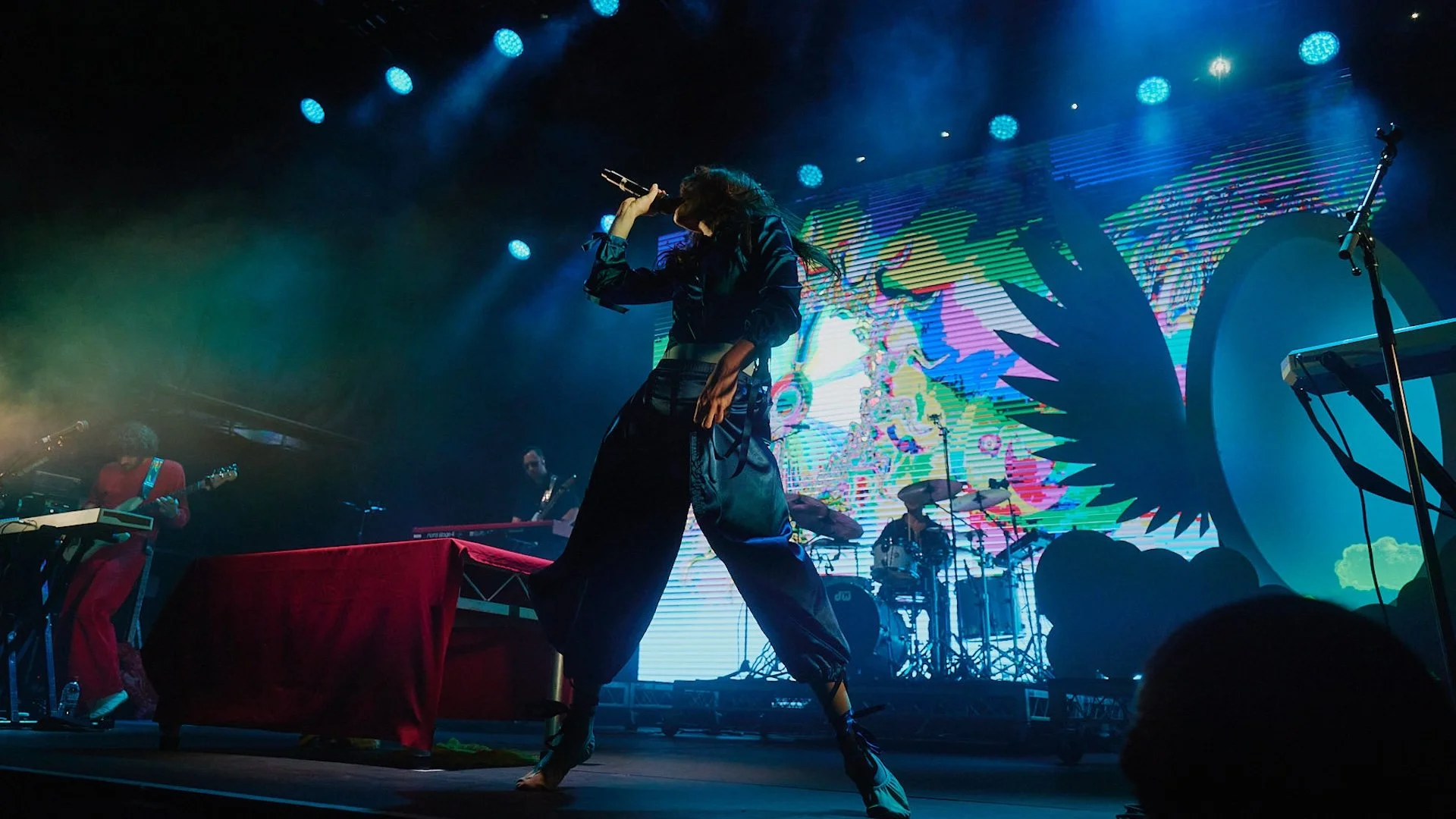 A female singer from Magdalena bay on stage at Palace Foreshore holding a microphone while performing with a band. The stage has colorful lighting and a digital background display.