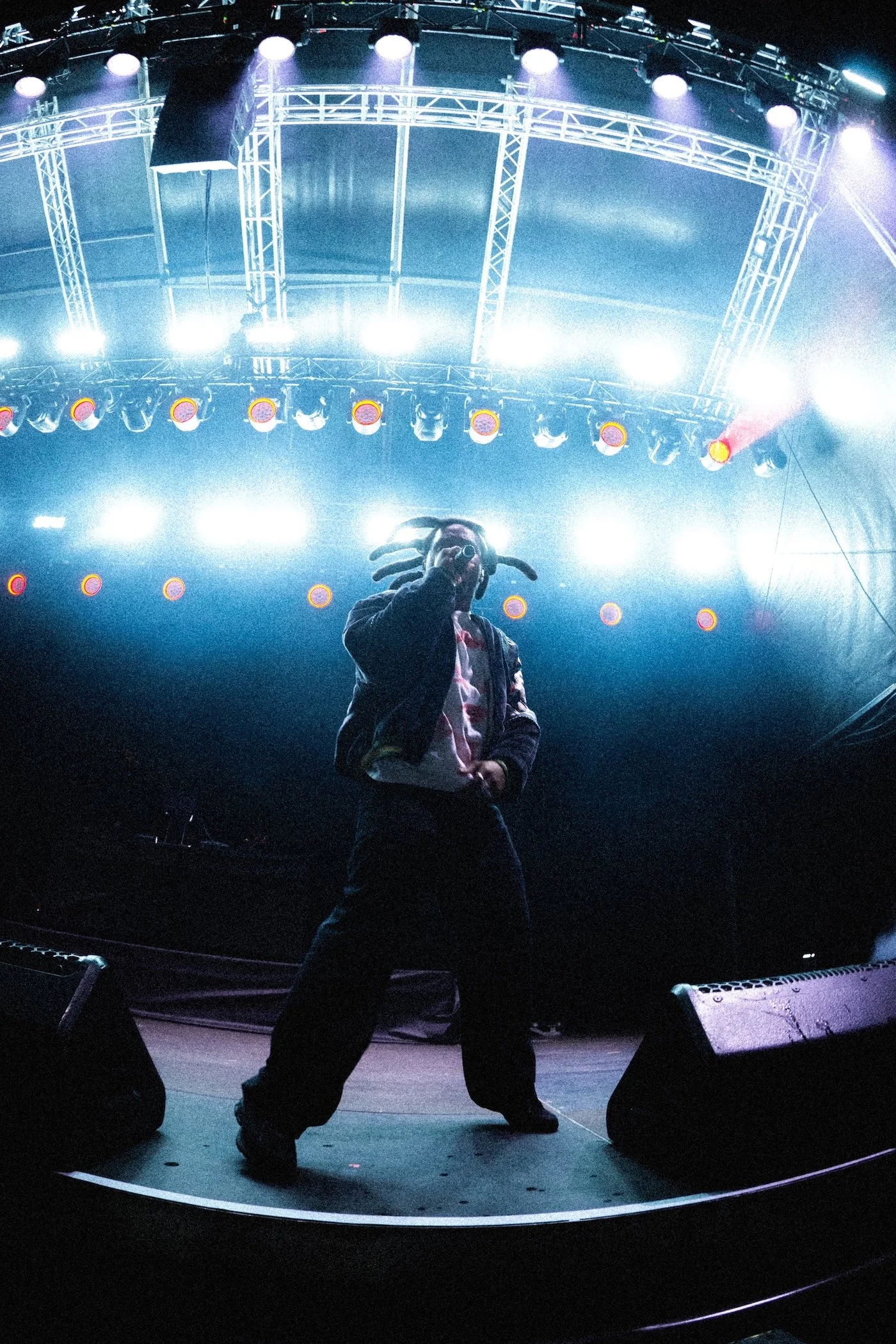 A performer with dreadlocks (Denzel Curry) wearing headphones and a jacket, holding a microphone, standing on a stage with bright lights and a metal truss overhead at a Palace Foreshore concert.