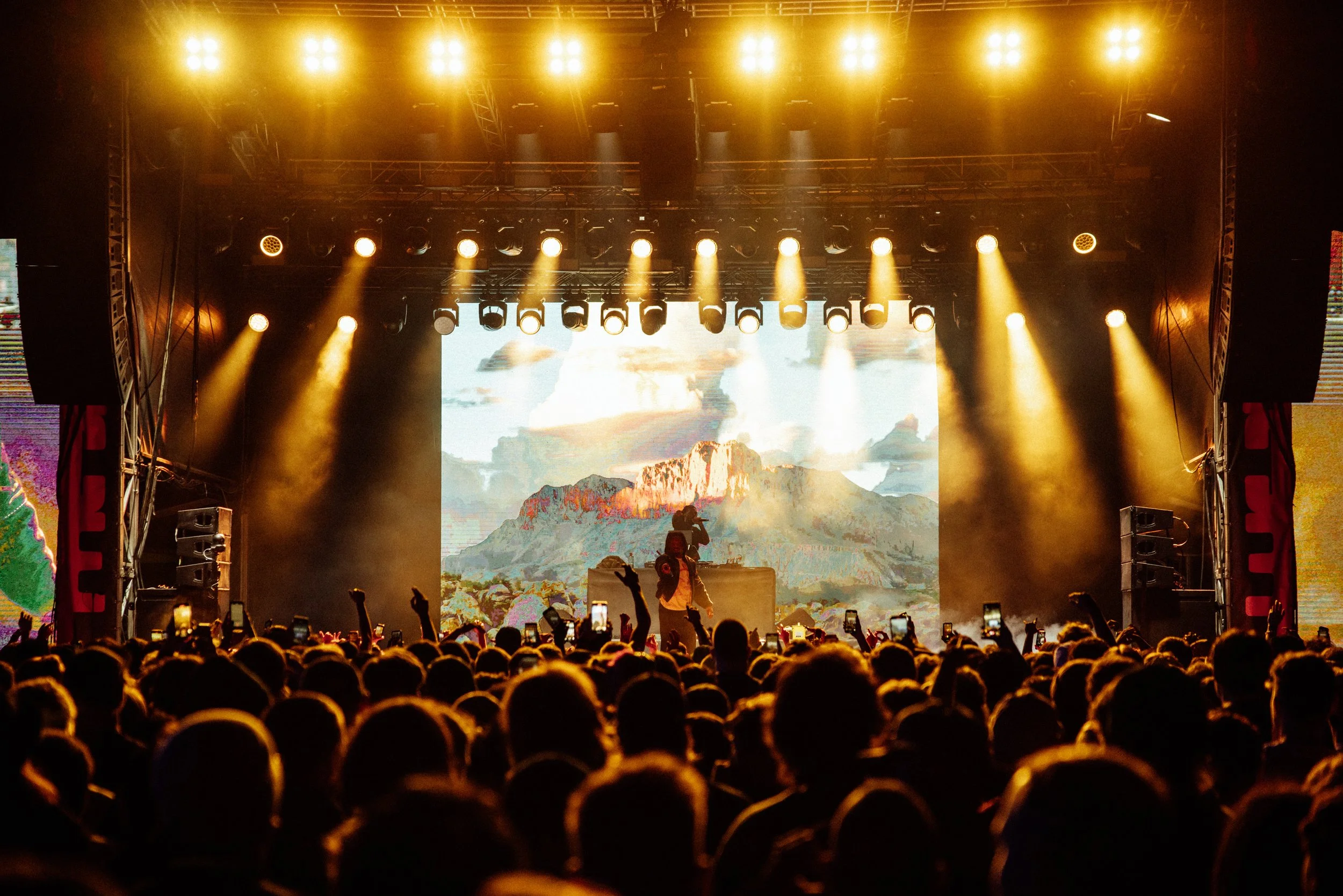 A crowded concert crowd watching Denzel Curry on stage at Palace Foreshore with a mountain landscape on a large screen behind them, surrounded by bright yellow stage lights.