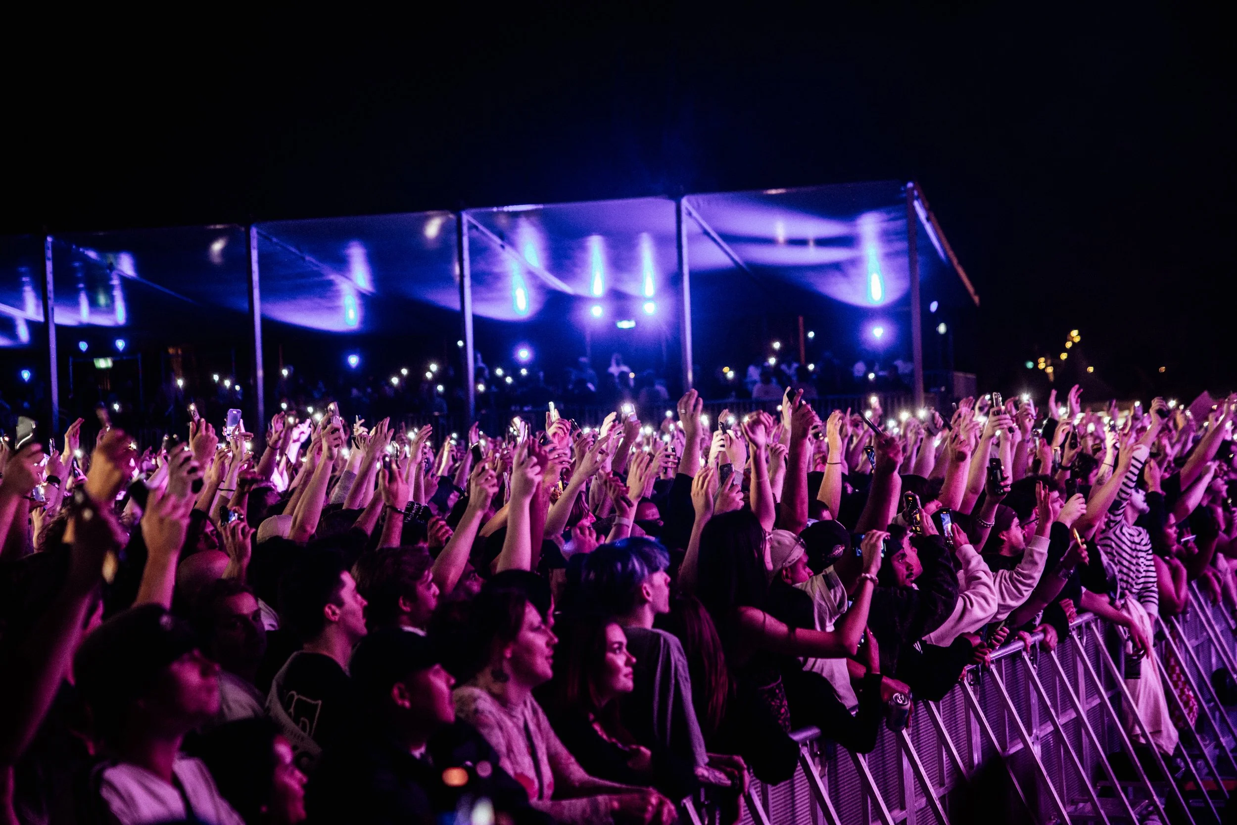 Crowd of concert attendees at Palace Foreshore raising their hands and taking photos at night, with stage lights visible in the background.
