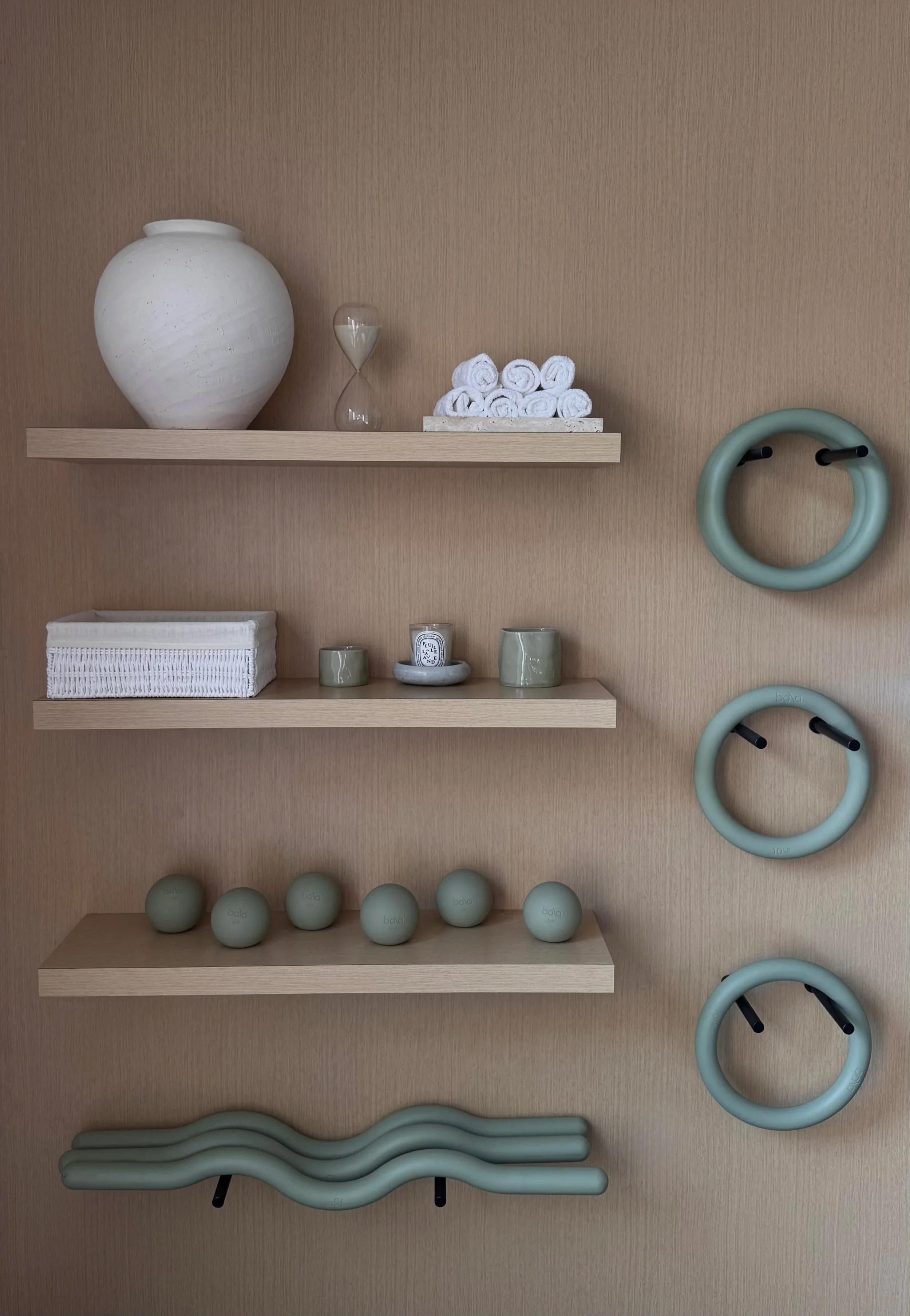 Estantes con objetos de gimnasio decorativos, incluyendo una vasija blanca, un reloj de arena, toallas enrolladas, pequeños recipientes, una vela y balones de ejercicio en color verde y una estructura de barras de gimnasia.