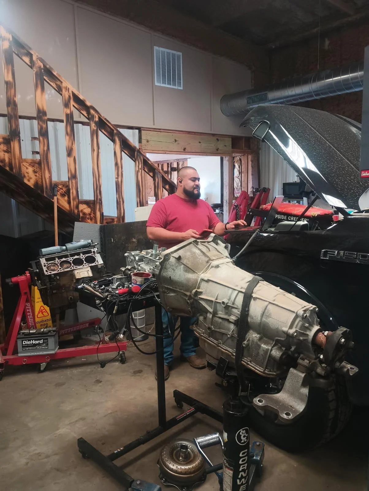 Man working on a car engine in a garage. The engine is attached to a transmission and situated on a stand. The man is focused, wearing a red shirt, and operating a laptop. The garage has a staircase and various tools around.