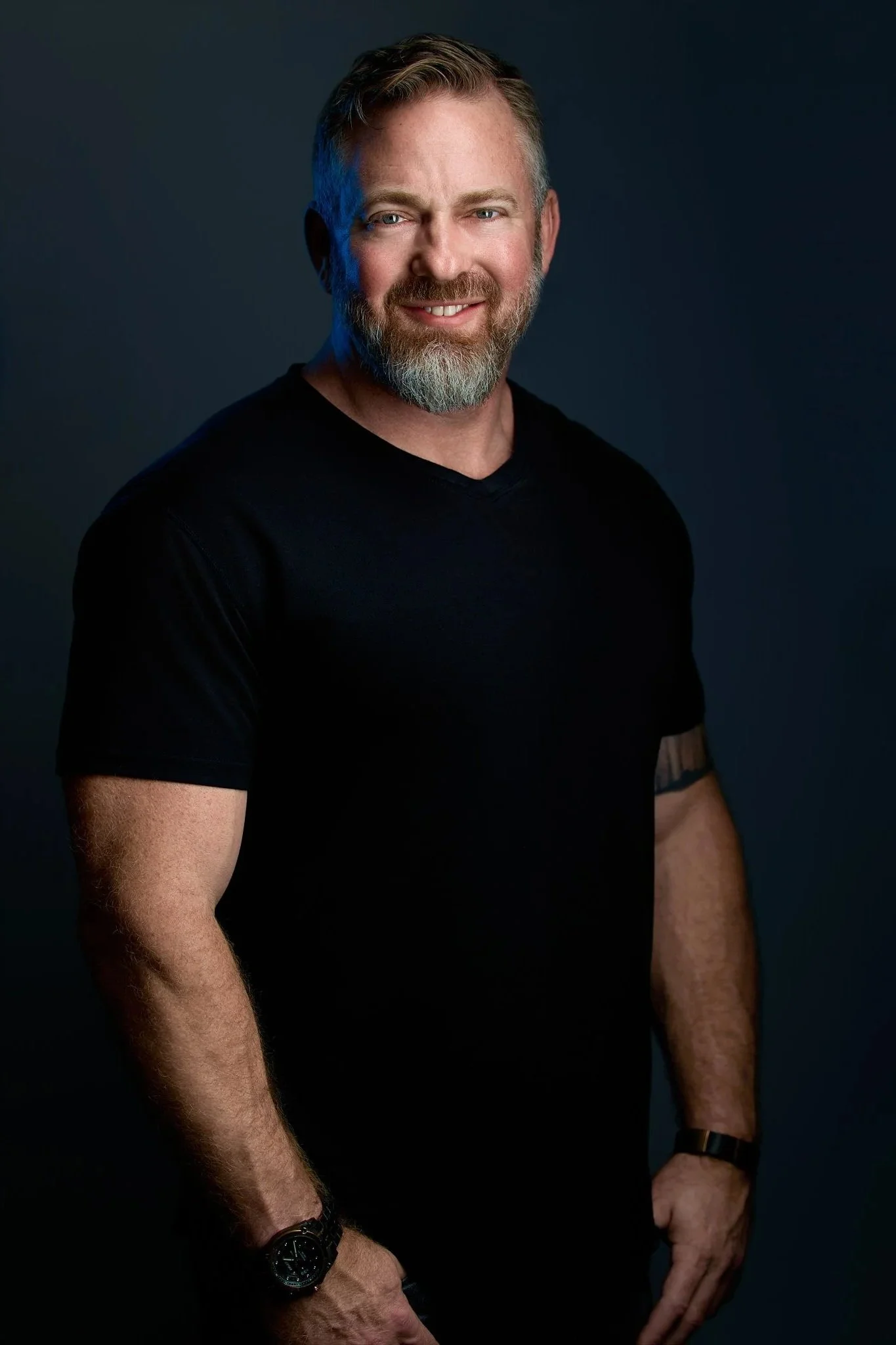 A smiling man with a beard, wearing a black t-shirt, standing against a dark background.