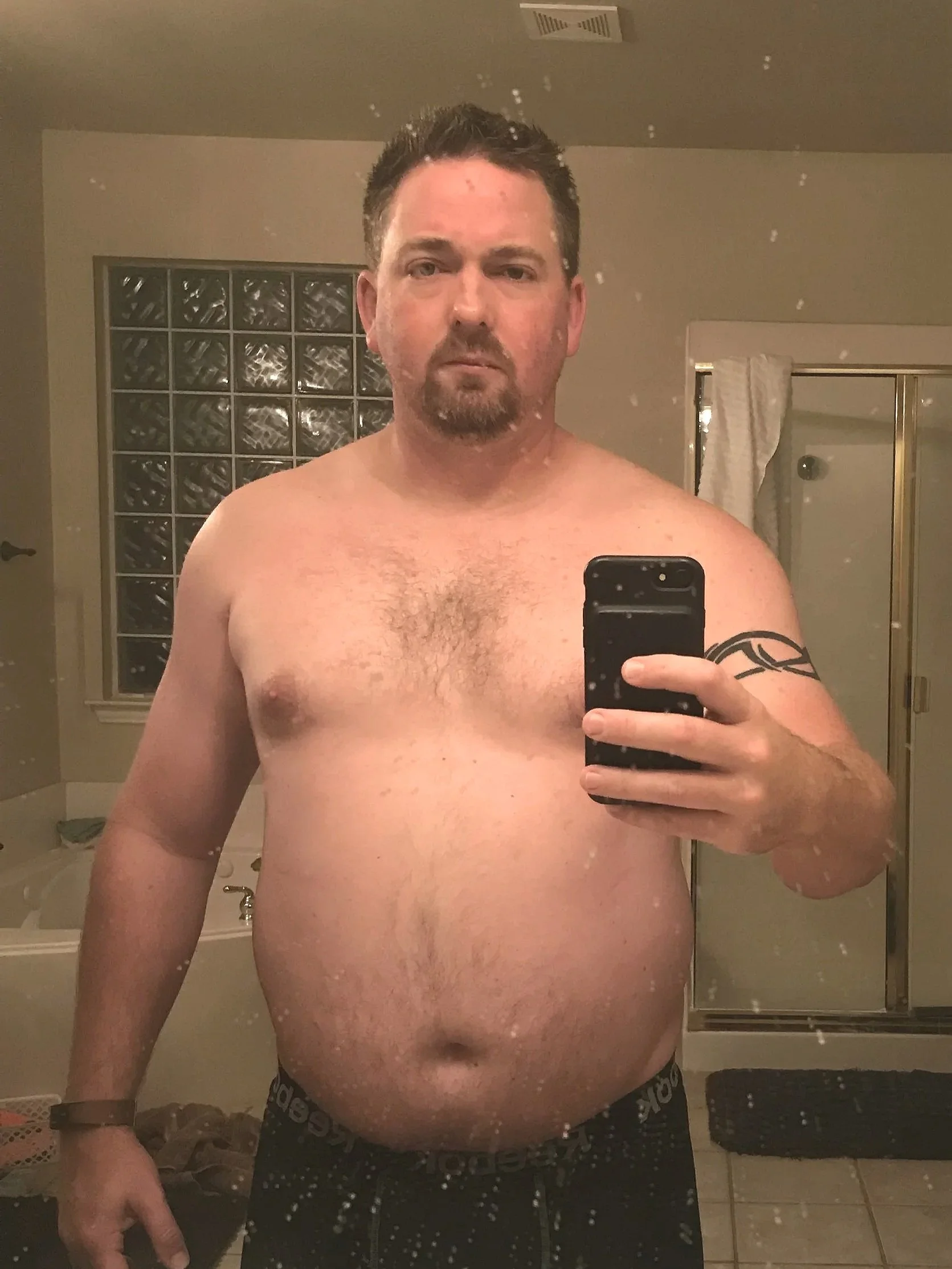 A shirtless man taking a mirror selfie in a bathroom, holding a smartphone in his right hand, posing for a before photo before beginning his fitness journey.