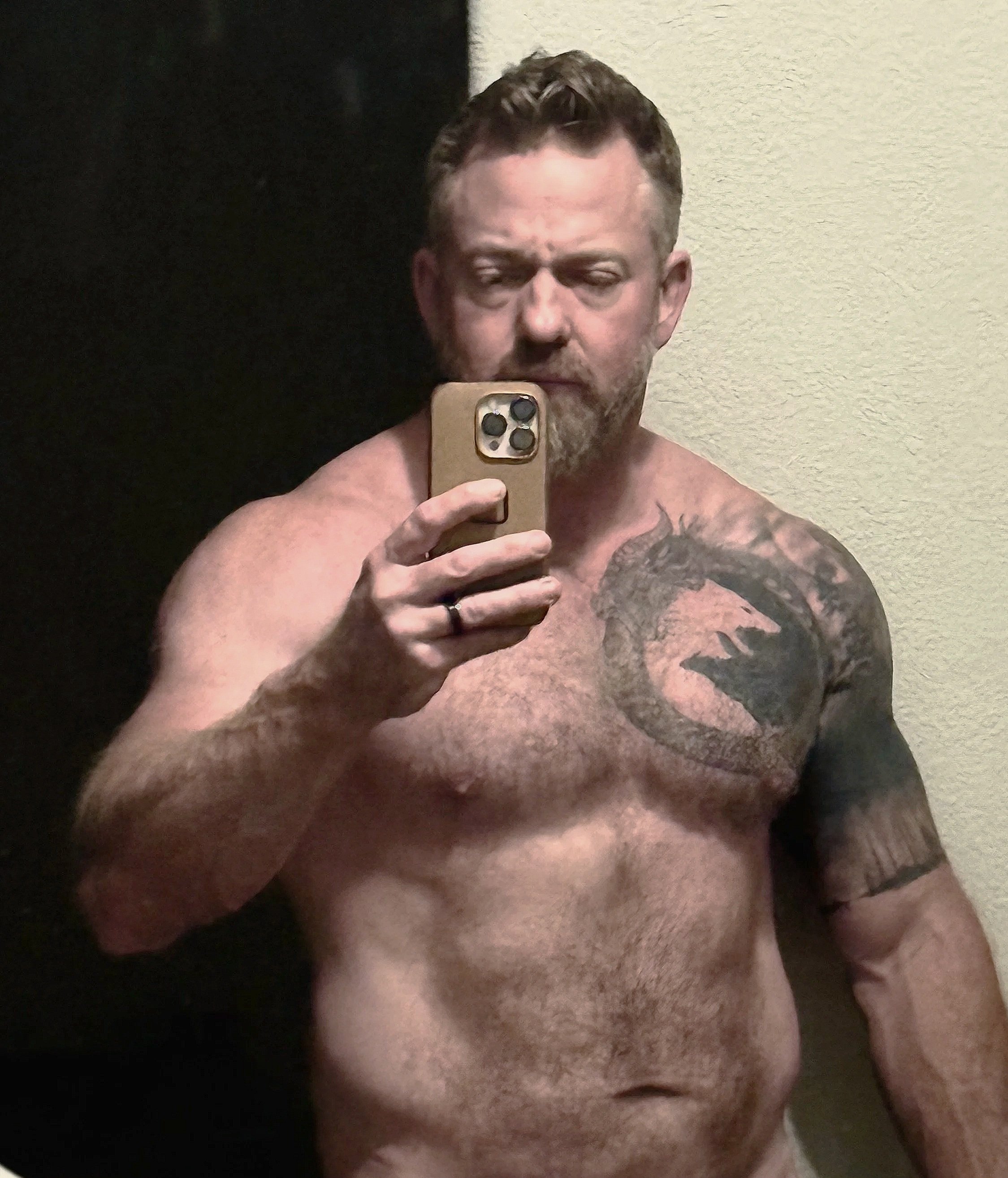 A shirtless man with a beard and short hair taking a mirror selfie with his phone posing in an after photo after a physical transformation.