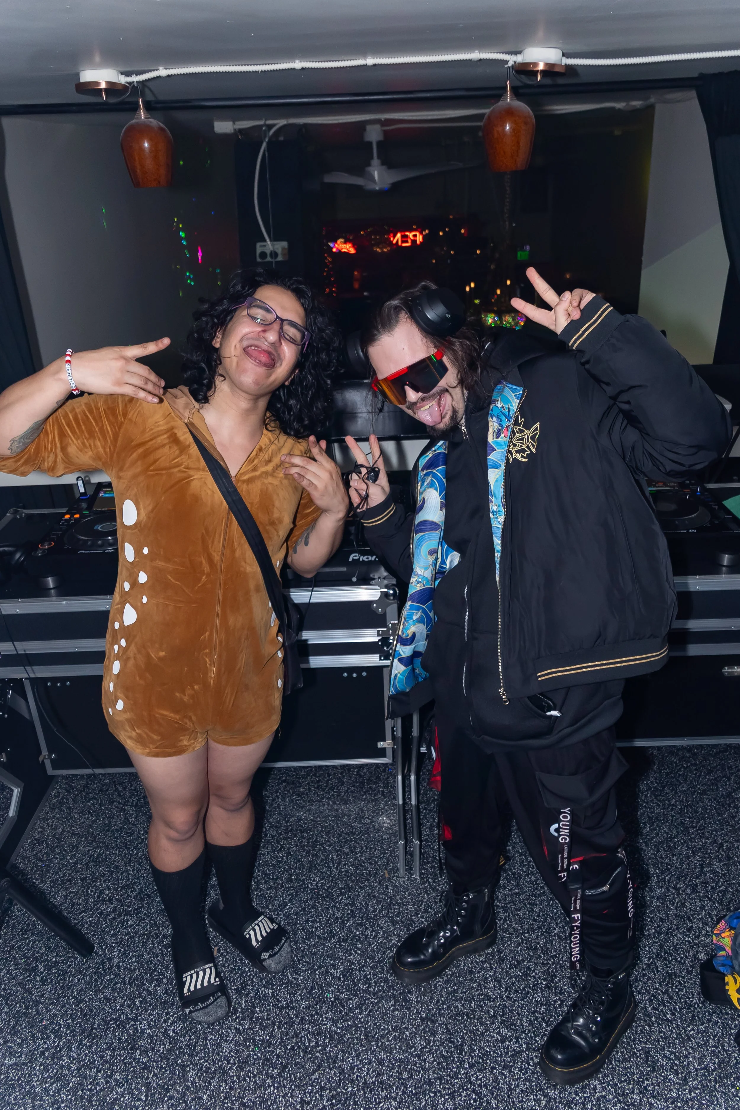 Two people dressed in casual and stylish clothing posing in a DJ booth with turntables, making peace signs and sticking out their tongues in a nightclub or party setting.