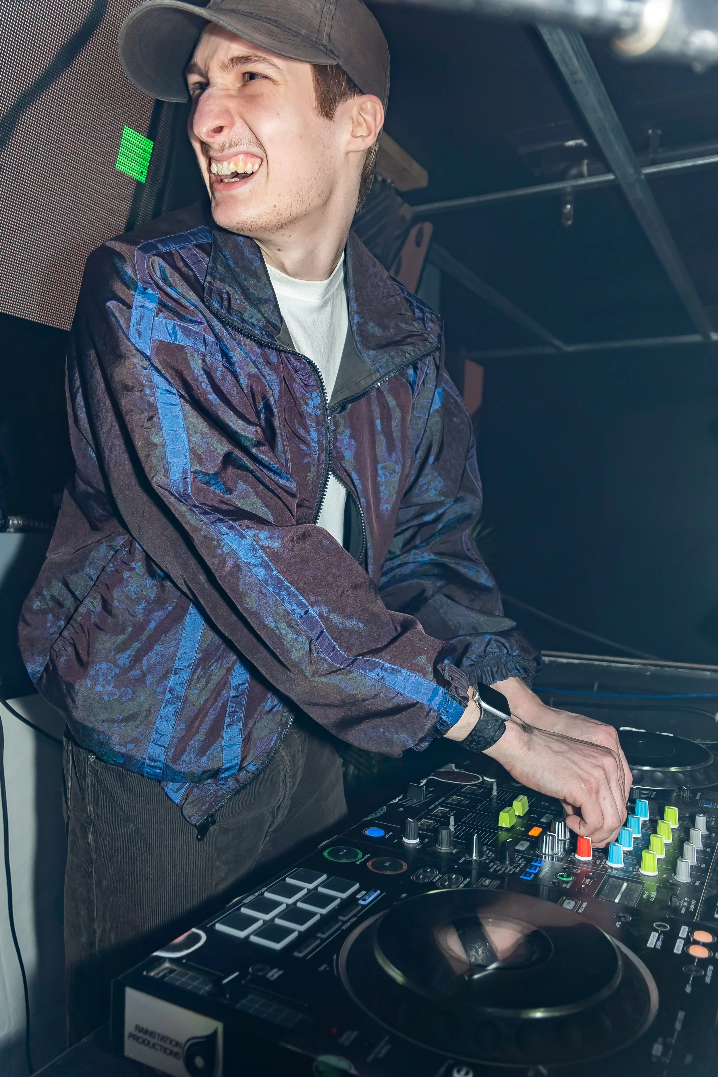 A DJ wearing a gray cap and a patterned jacket, smiling and operating a DJ mixer and turntables in a dimly lit setting.