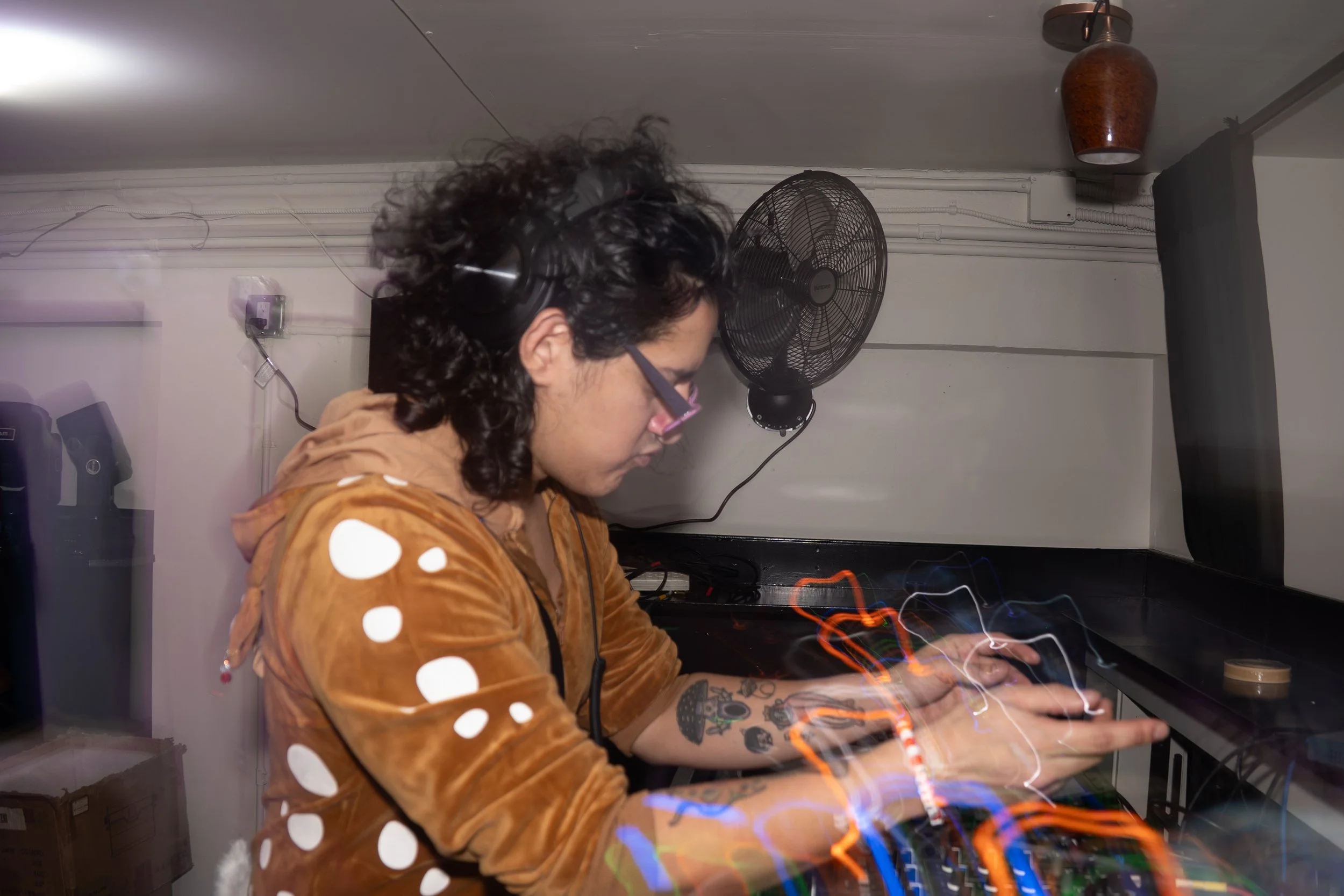 Person with curly hair, glasses, headphones, and tattoos wearing a brown polka-dot hoodie working with electronic music equipment in a small room.