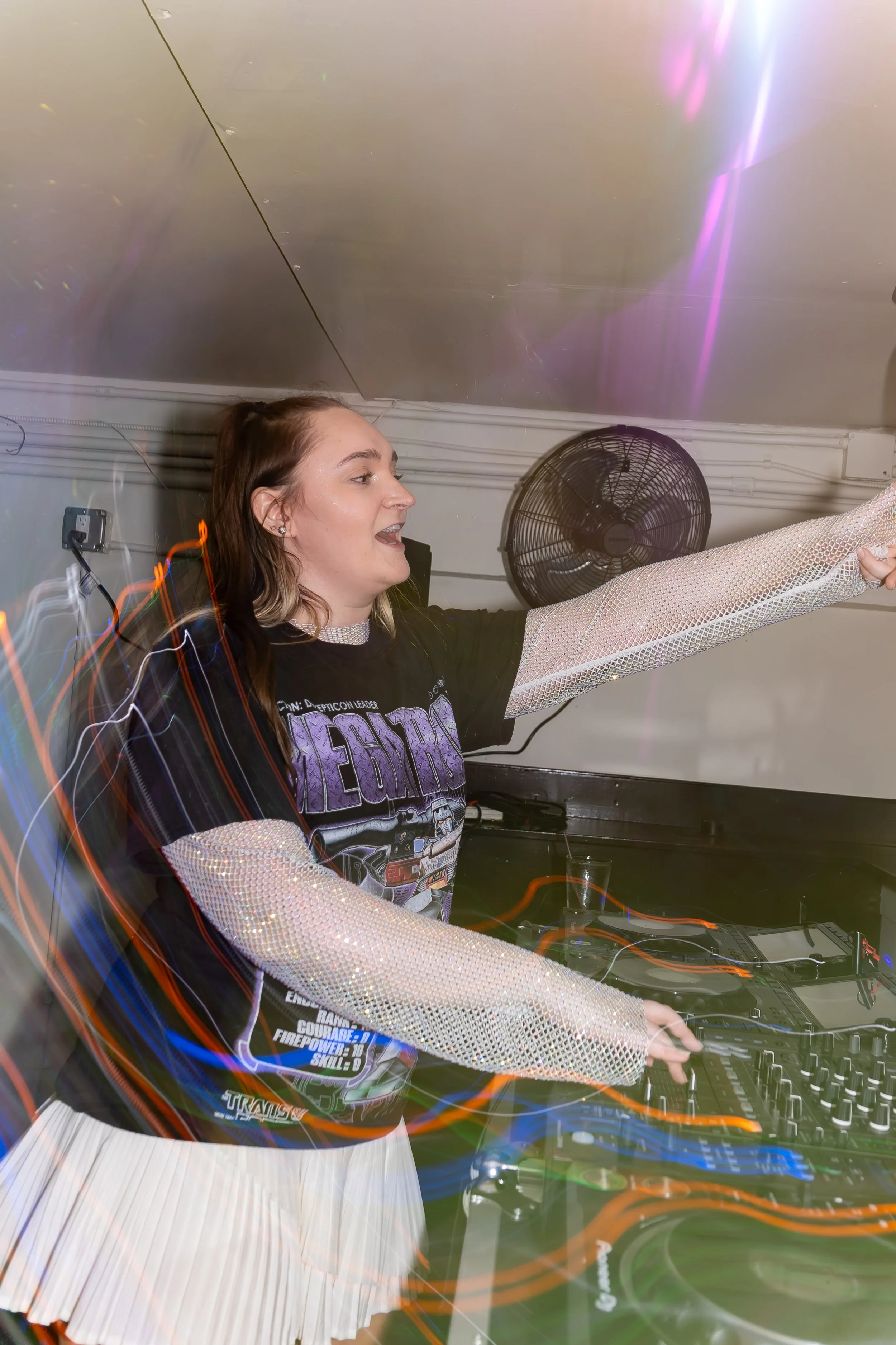 A woman DJ wearing a black T-shirt with a graphic and a white pleated skirt, with her arm raised pointing, mixing music at a party with colorful light streaks and a fan in the background.