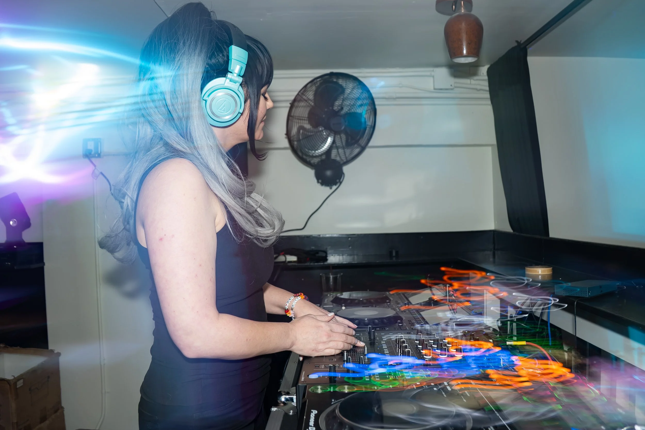 A woman with long, colored hair wearing headphones is DJing at a music event or party, with DJ equipment and colorful light effects.