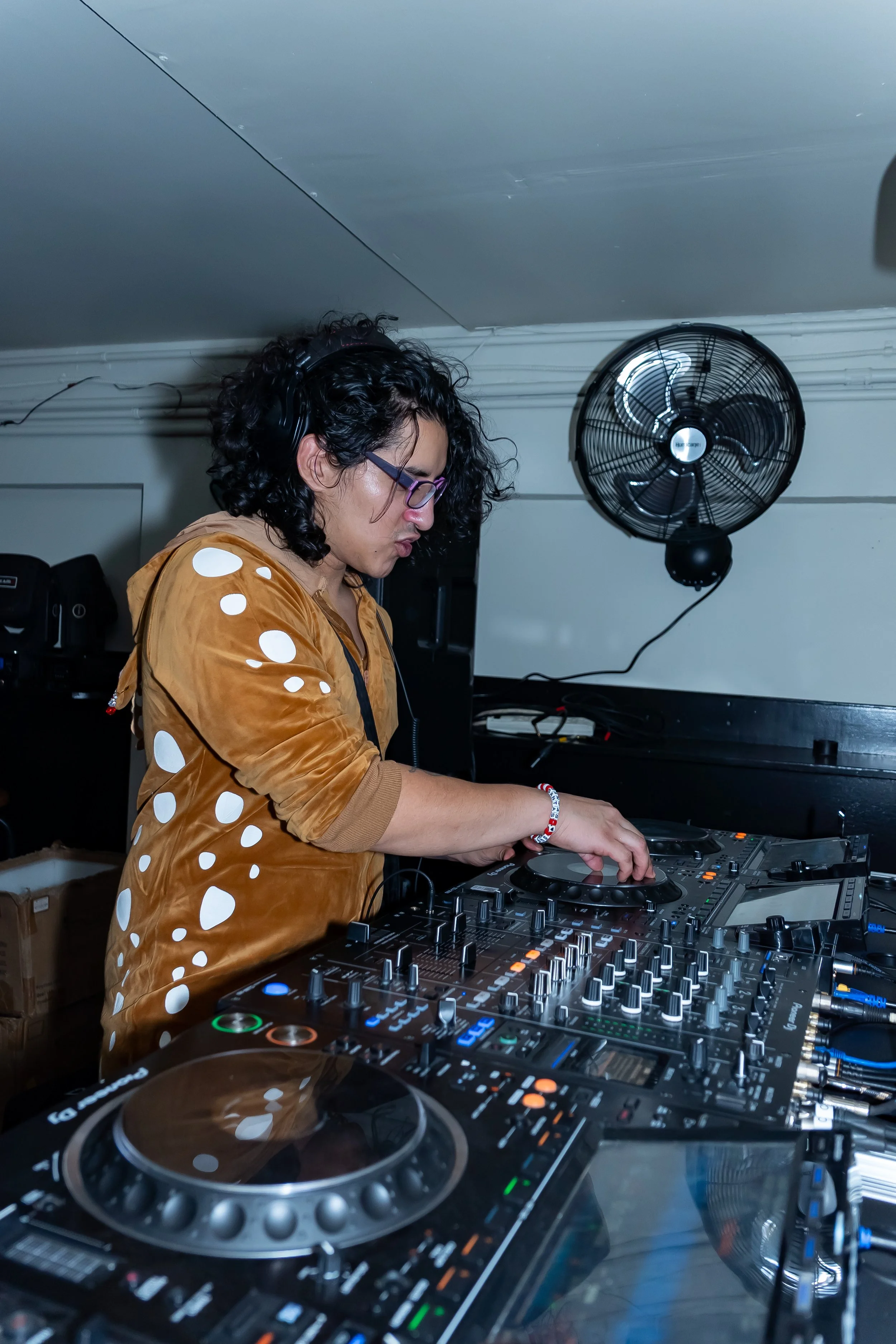 A DJ with curly black hair and glasses, wearing a tan jacket with white polka dots, is mixing music on professional DJ equipment. A black ceiling fan is mounted on the wall behind her.