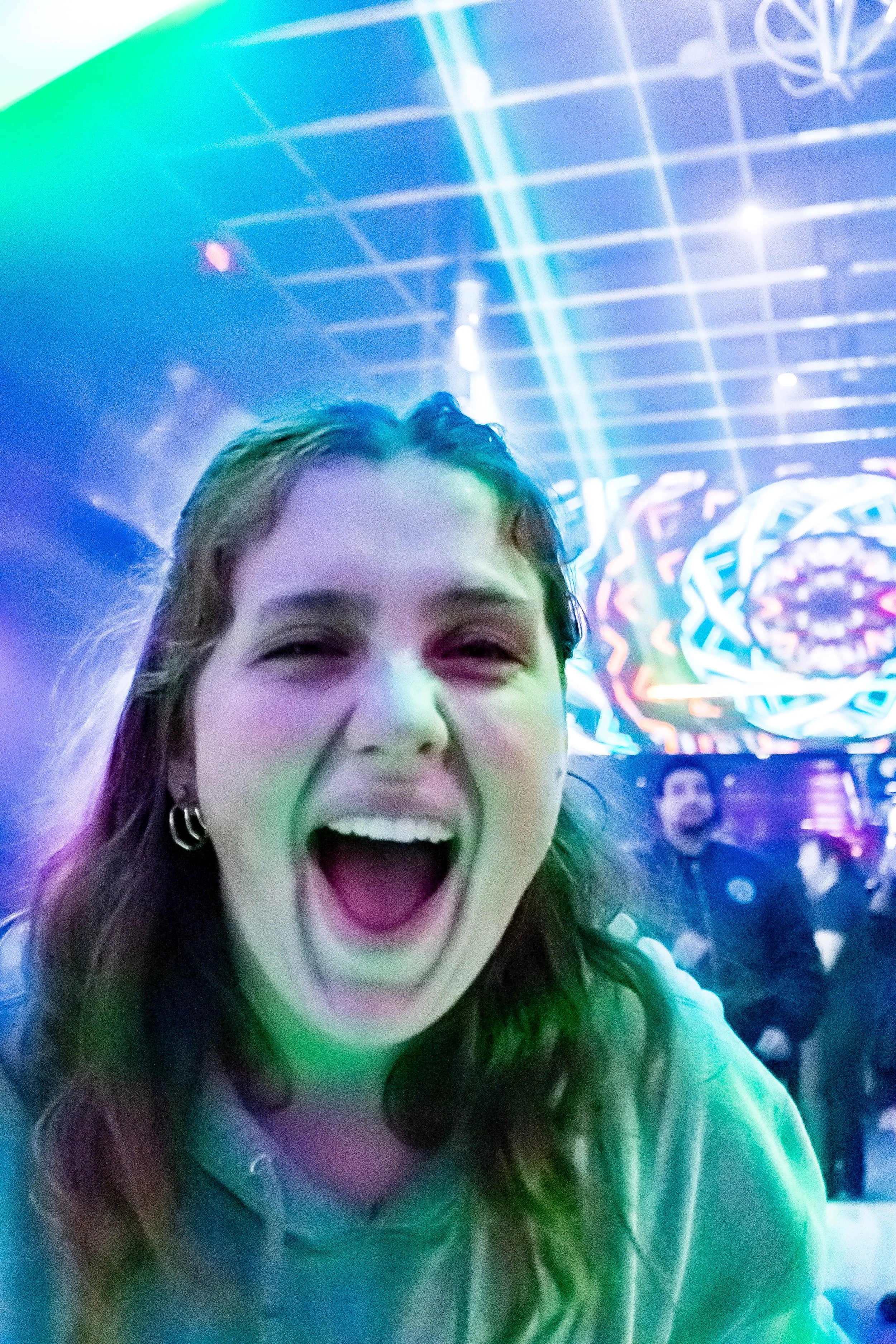 Close-up of young woman with long hair smiling widely and shouting at a vibrant light show or concert with neon visuals and other people in the background.