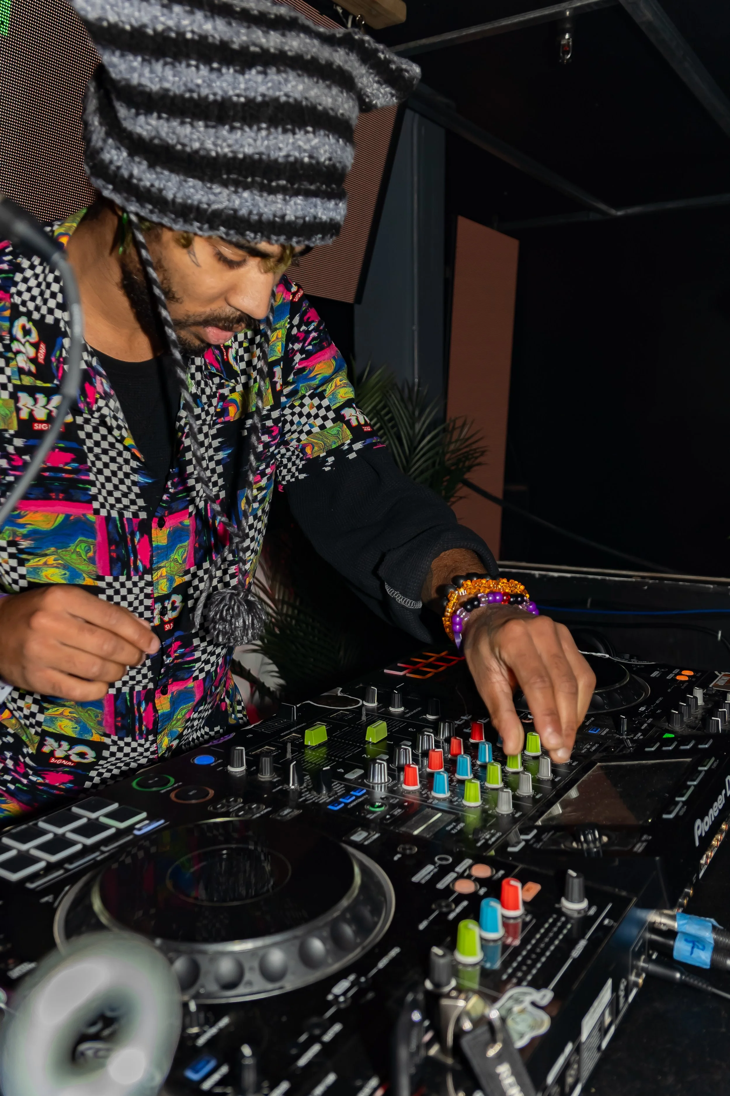 A DJ wearing a colorful, patterned shirt and a black and white striped hat, mixing music on a Pioneer DJ controller in a dark indoor venue.
