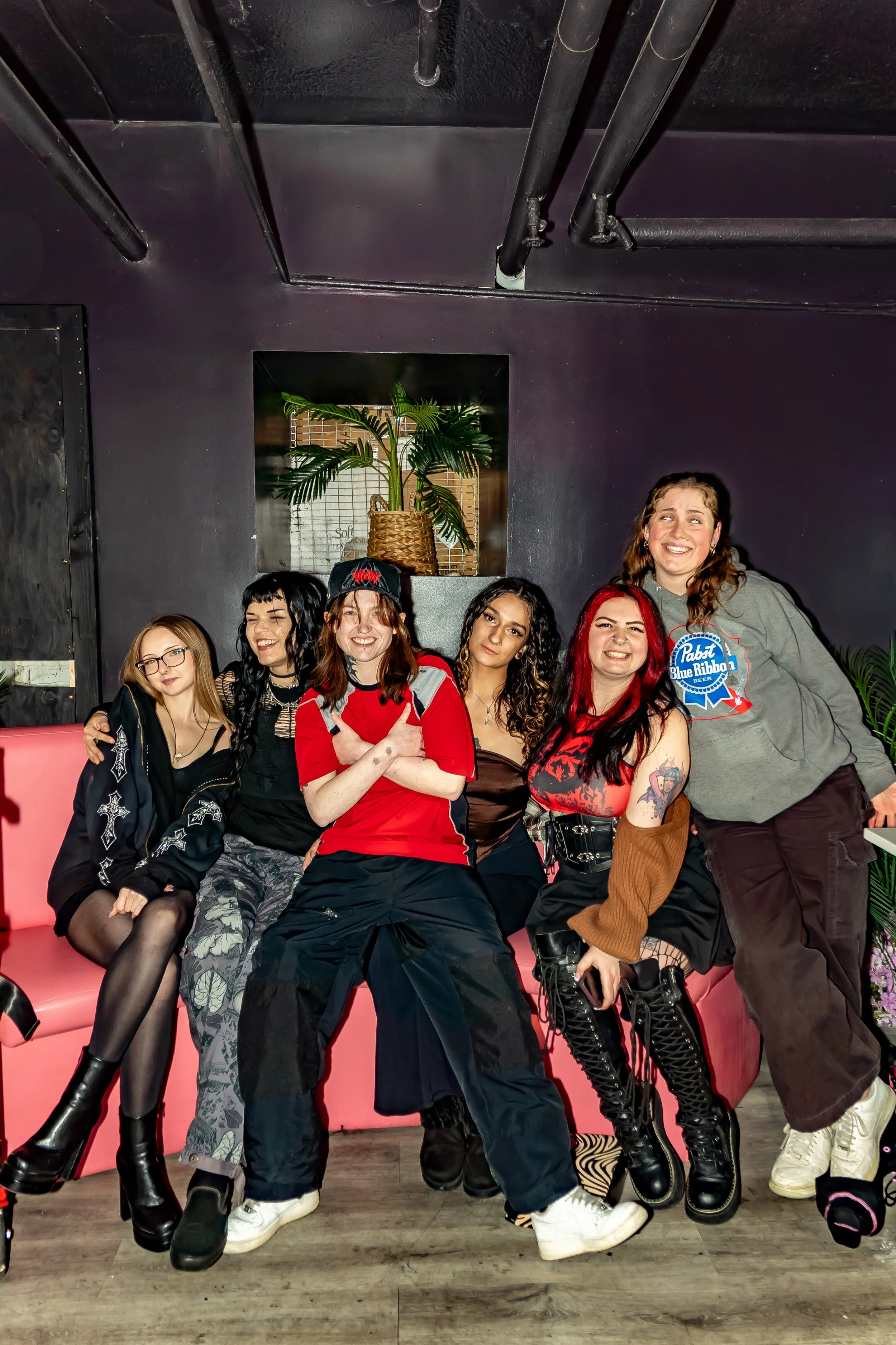 Group of six young women sitting on a pink bench in a room with dark walls, posing and smiling for a photo. There are plants and a black wall in the background.