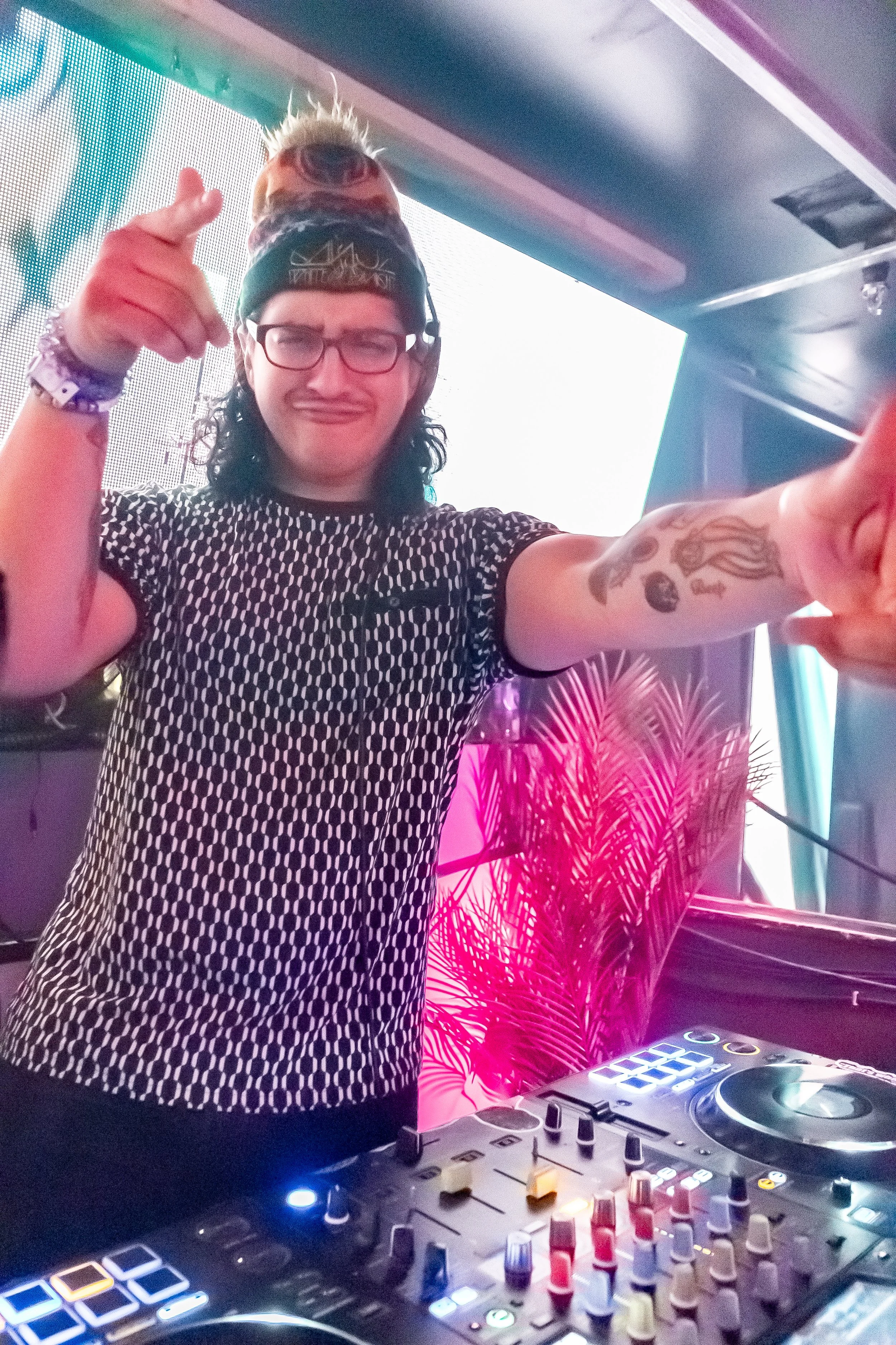 A DJ with glasses, tattoos, and long dark hair is standing behind a DJ controller, pointing towards the camera, in a colorful club or event space with bright lights and tropical plant decor.