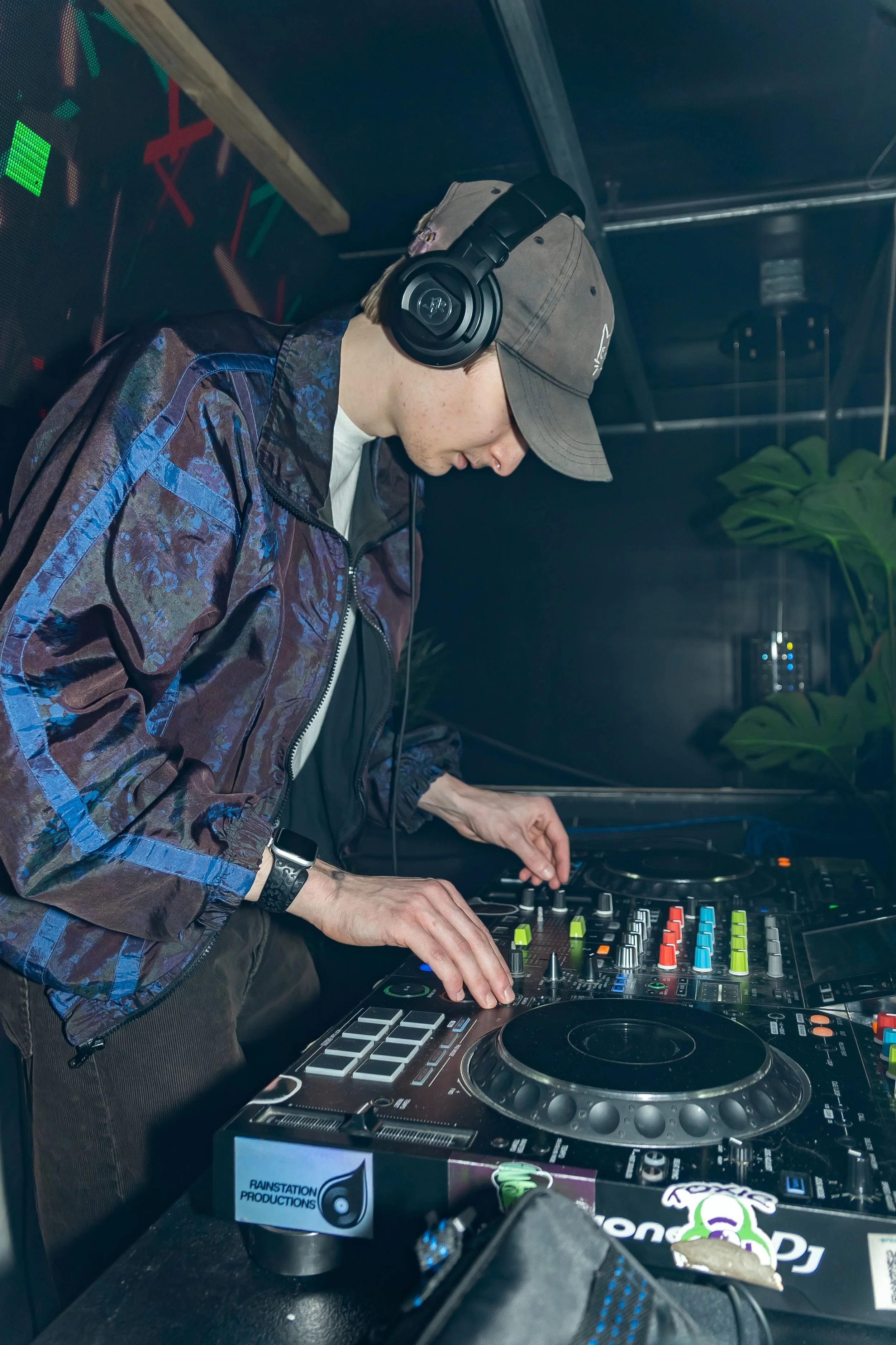 A DJ wearing headphones, a gray cap, and a patterned jacket, mixing music on a professional DJ controller in a dimly lit setting, possibly a club or event.