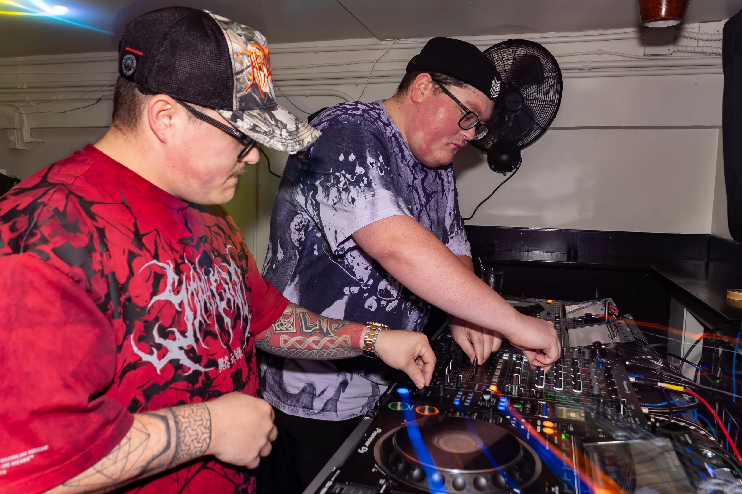 Two men DJing with a DJ controller, one wearing a red tie-dye shirt and black cap, the other in a purple tie-dye shirt and black beanie, in a club or party setting.