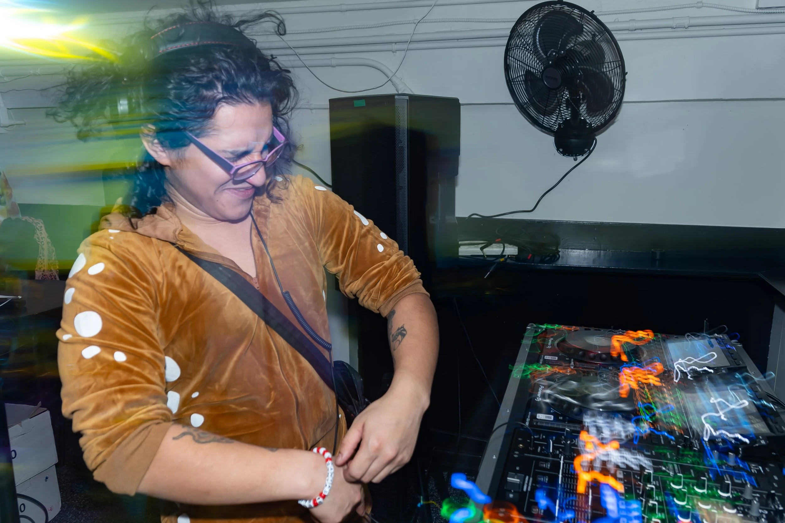 A person with curly hair, glasses, and tattoos smiling while DJing at a music event with colorful lights and equipment.