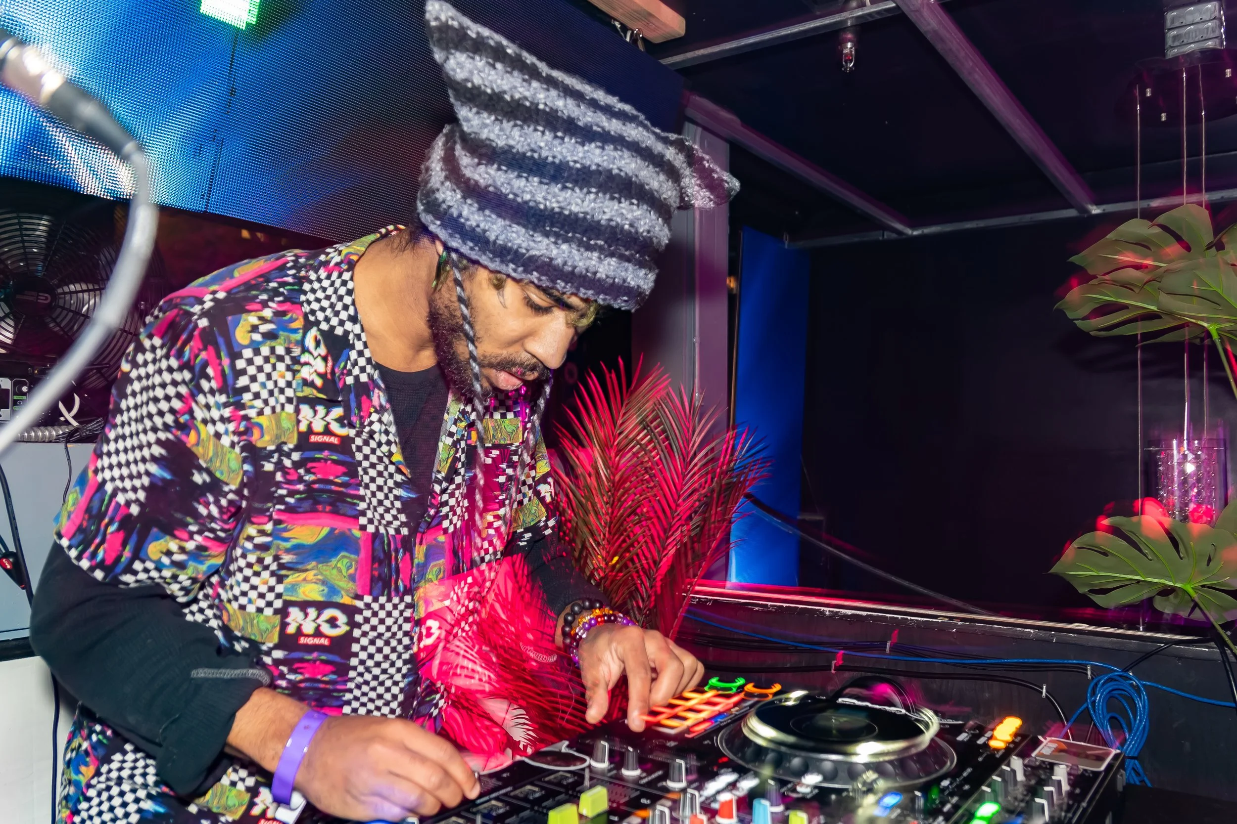 A DJ is performing at a nightclub with colorful lighting and tropical plants in the background.