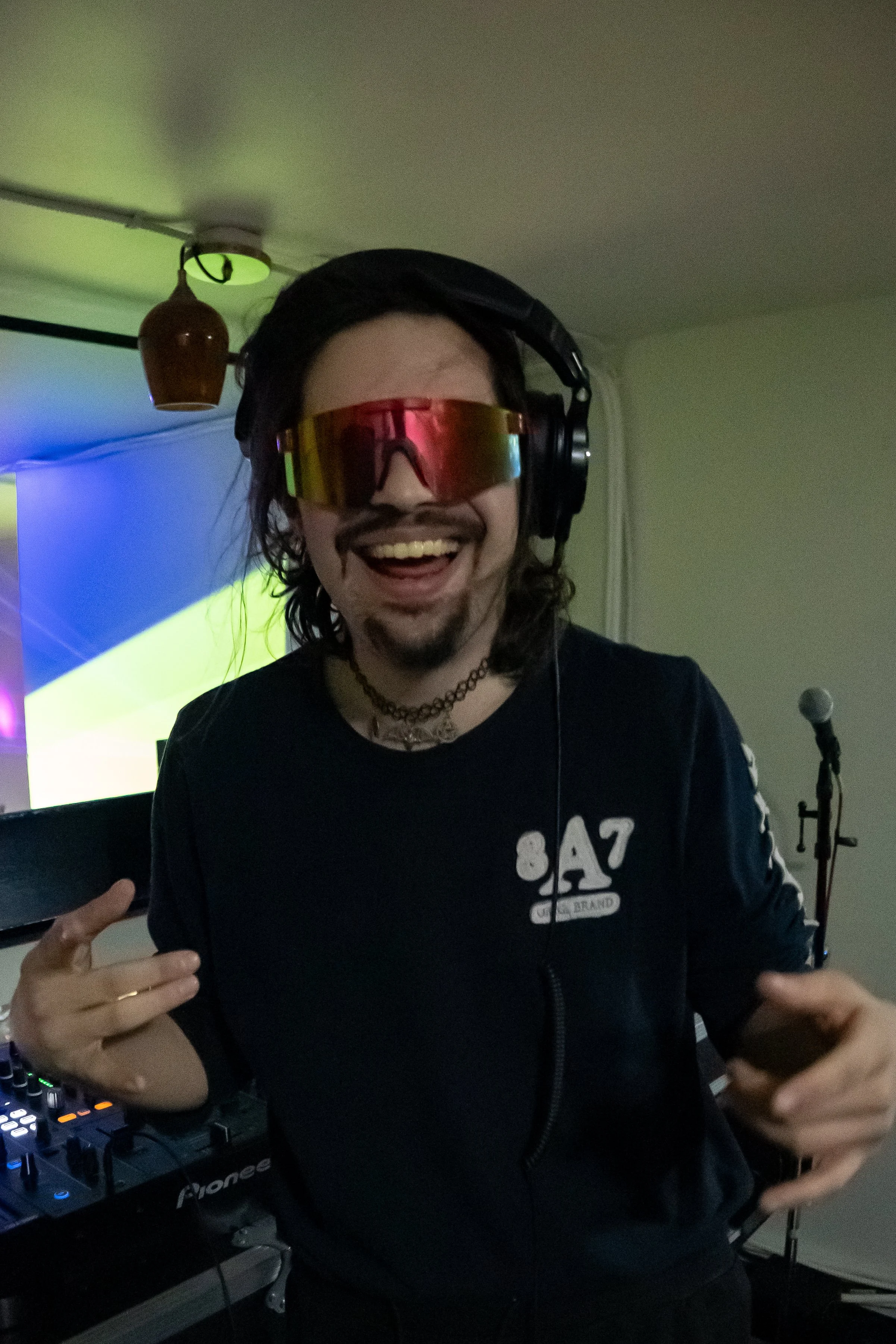 A person with long hair wearing reflective sunglasses, headphones, a black jacket, and a choker, standing by DJ equipment in a room with a microphone, smiling and making a gesture with their hands.