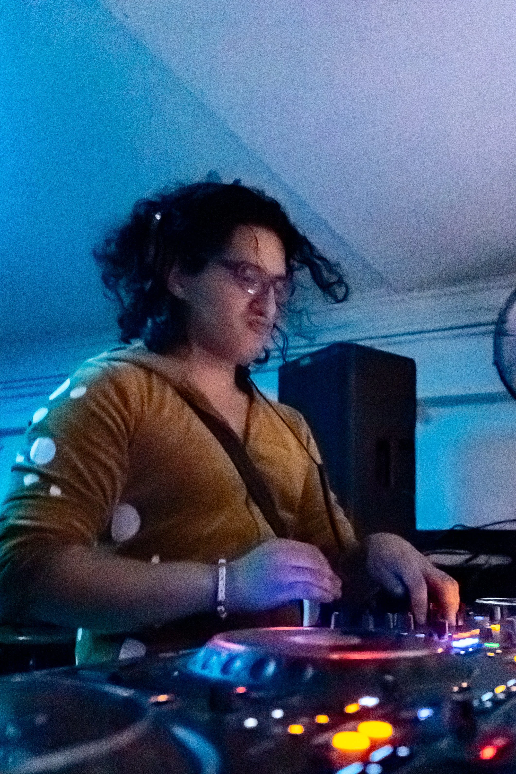 A woman with curly hair and glasses DJing at a nightclub under blue lighting.