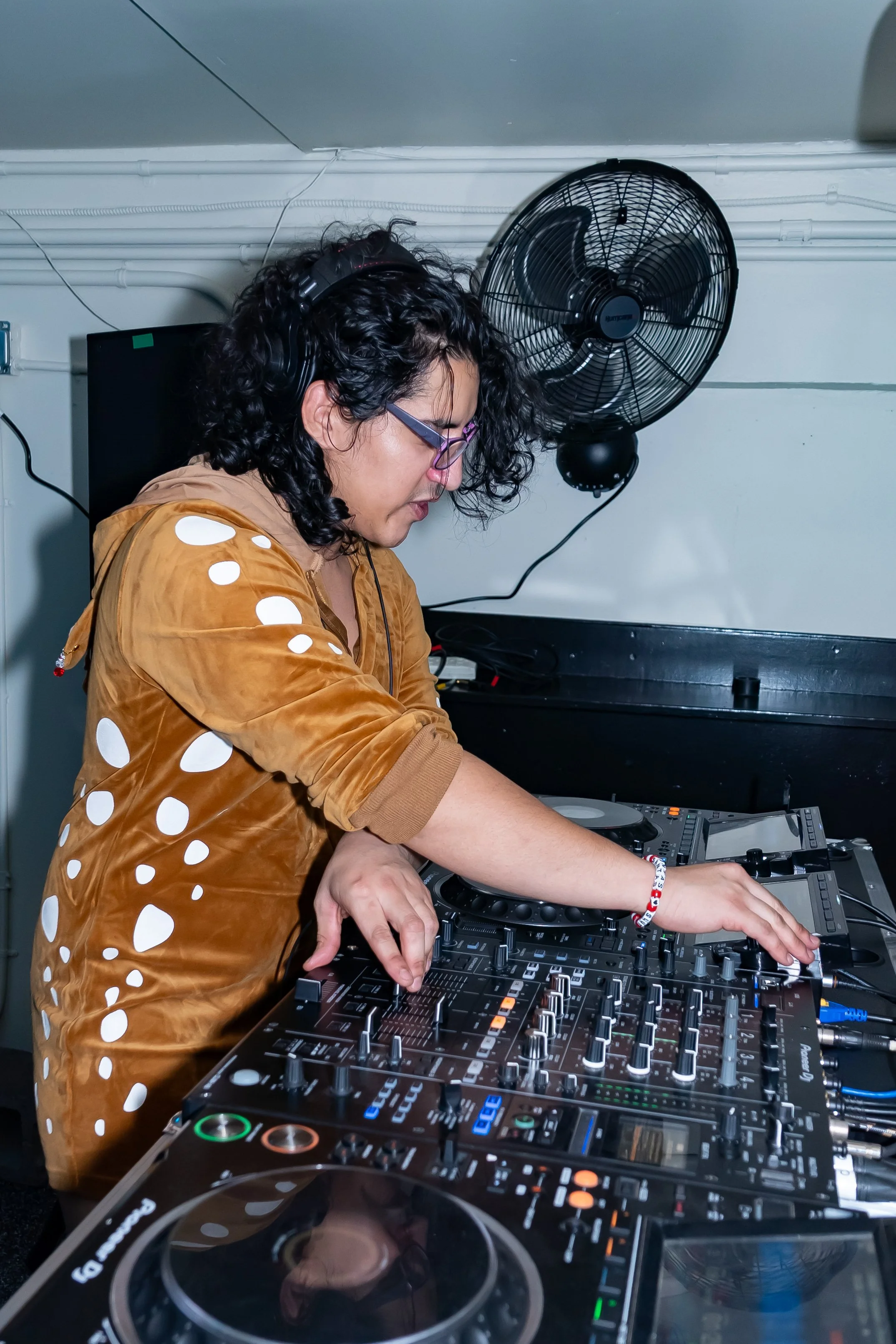 A female DJ wearing glasses and a brown jacket with white polka dots, DJing with turntables and a mixer in a room with a wall-mounted fan.