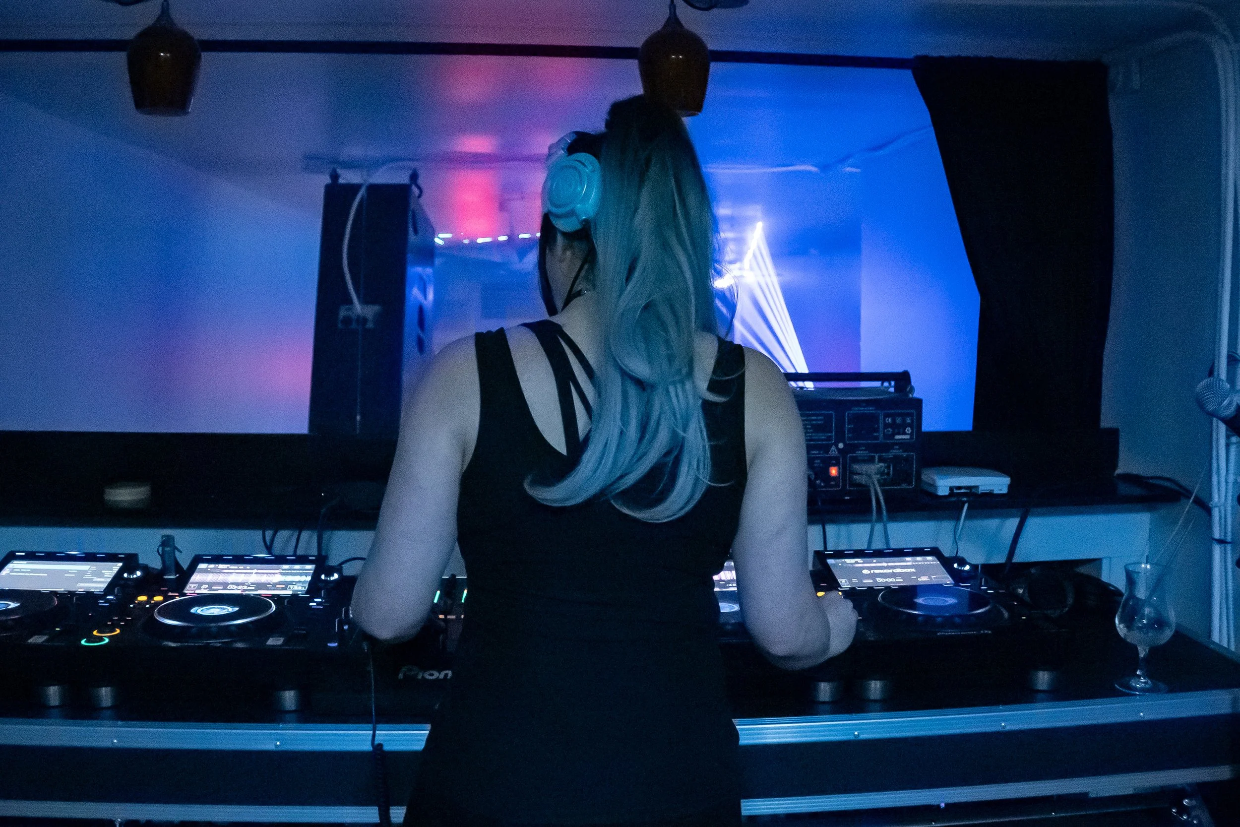 A female DJ with long, colored hair and headphones mixing music at a club or party with blue and purple lighting.