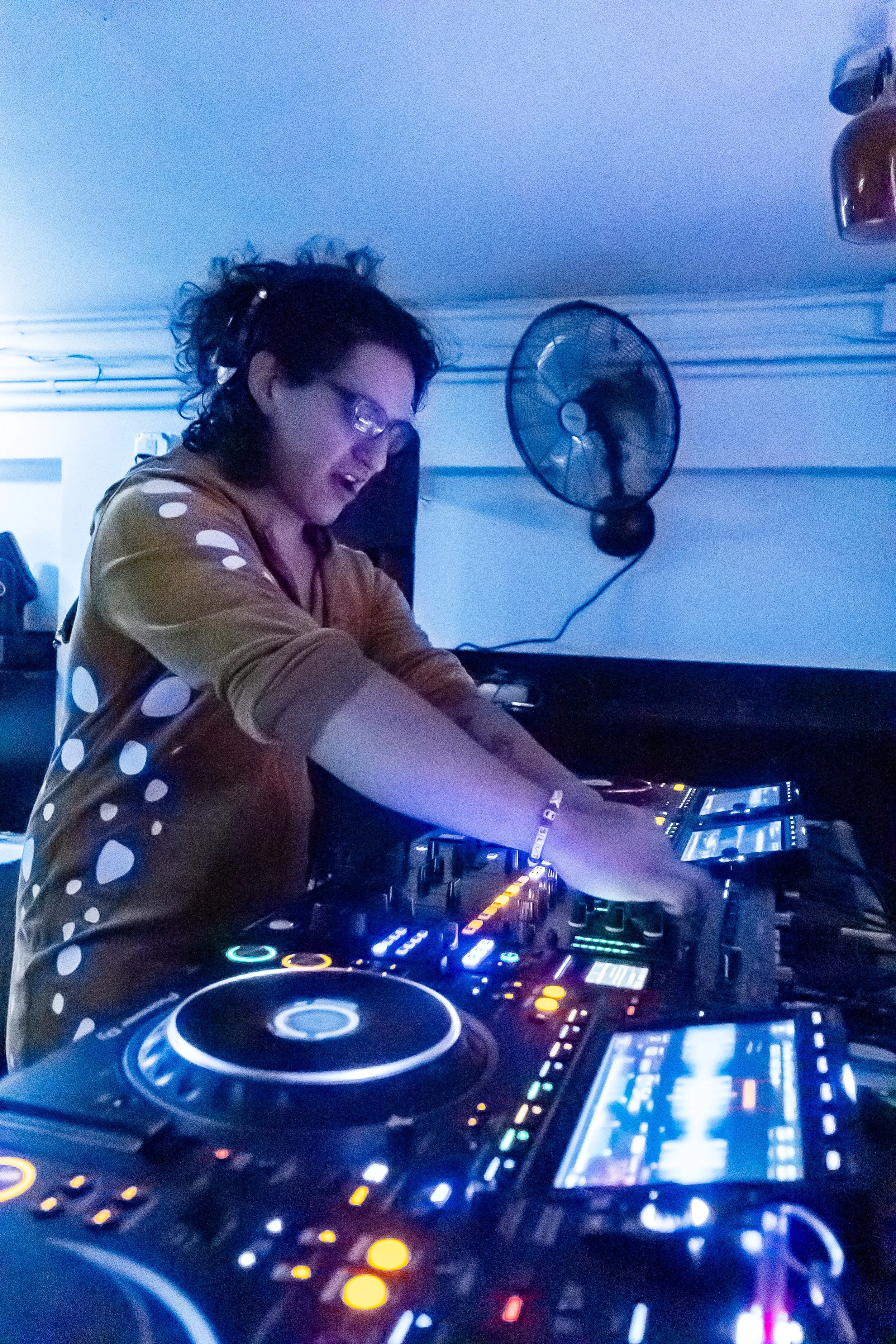 A DJ wearing glasses and a polka dot shirt mixes music on a DJ controller with colorful lights, in a dimly-lit setting with a fan on the wall.