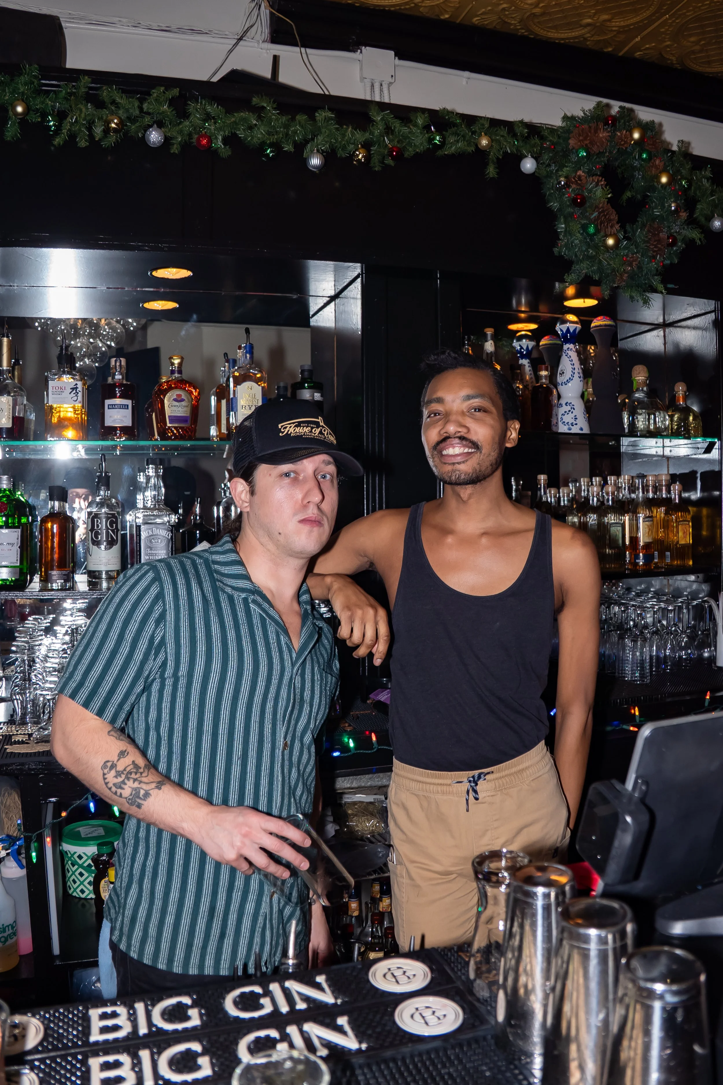 Two men behind a bar counter with various liquor bottles and glasses, one in a striped shirt and cap, the other in a black tank top, with Christmas decorations hanging above.