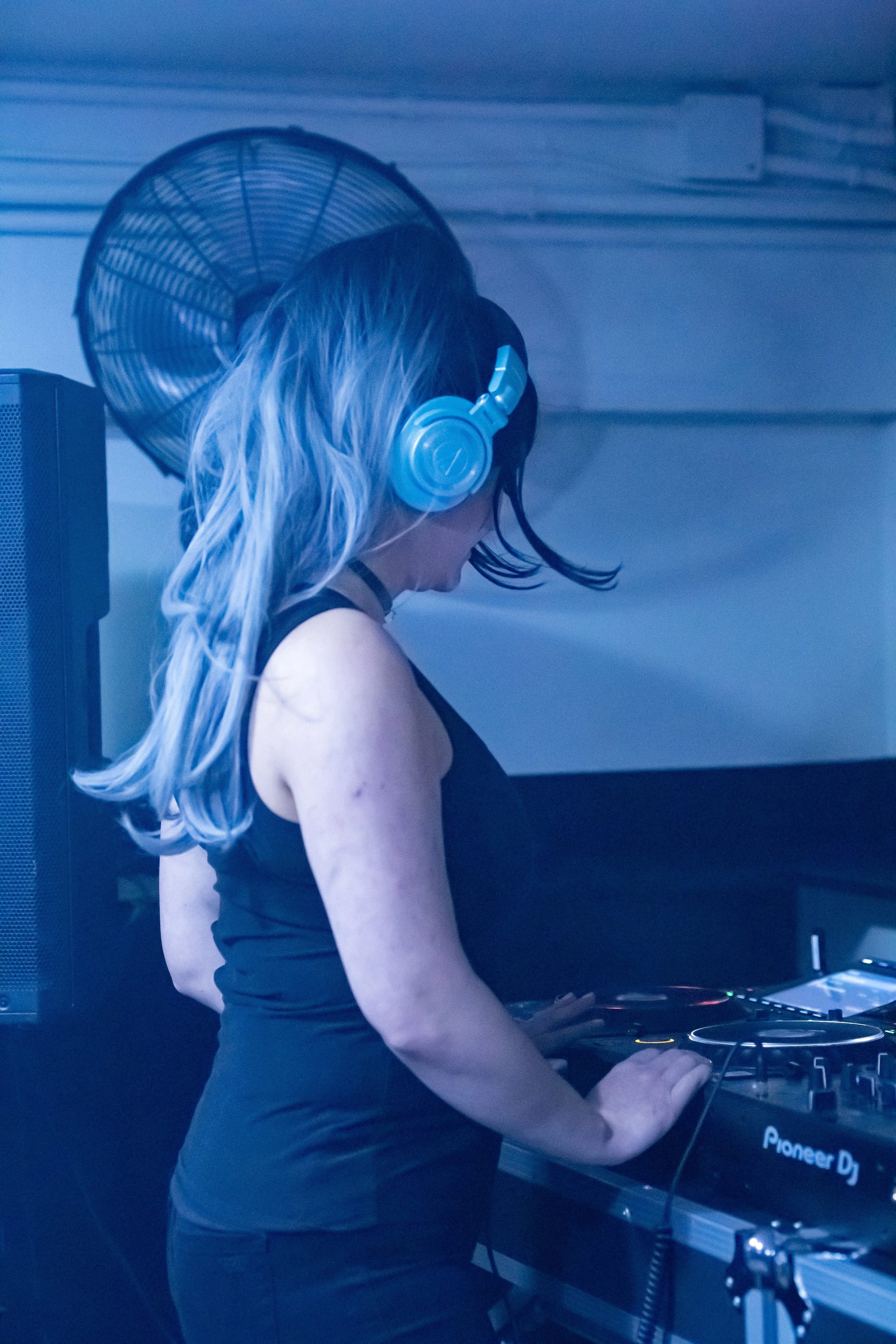 A woman with long, wavy blonde hair wearing a black sleeveless top and headphones, DJing at a mixing console in a dimly lit room.